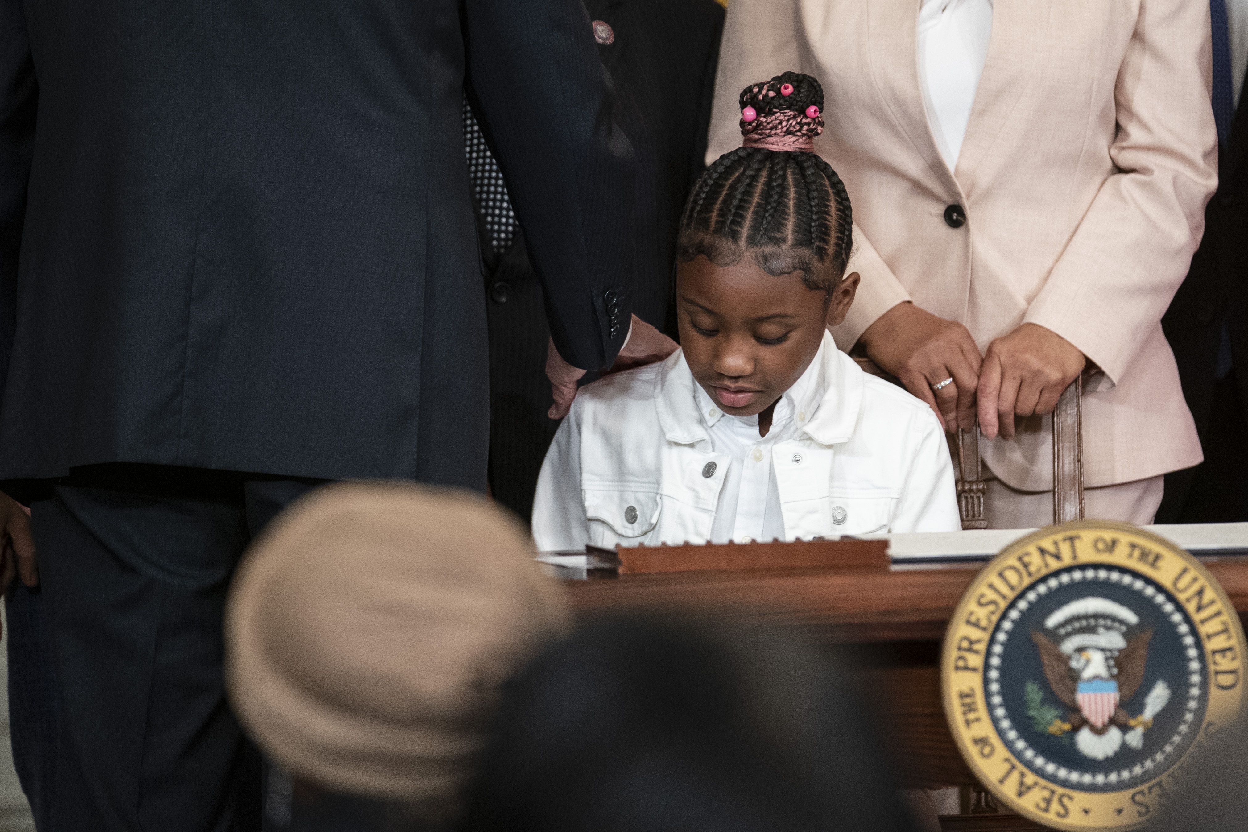 Photo of a young Black girl looking down as people crowd behind her