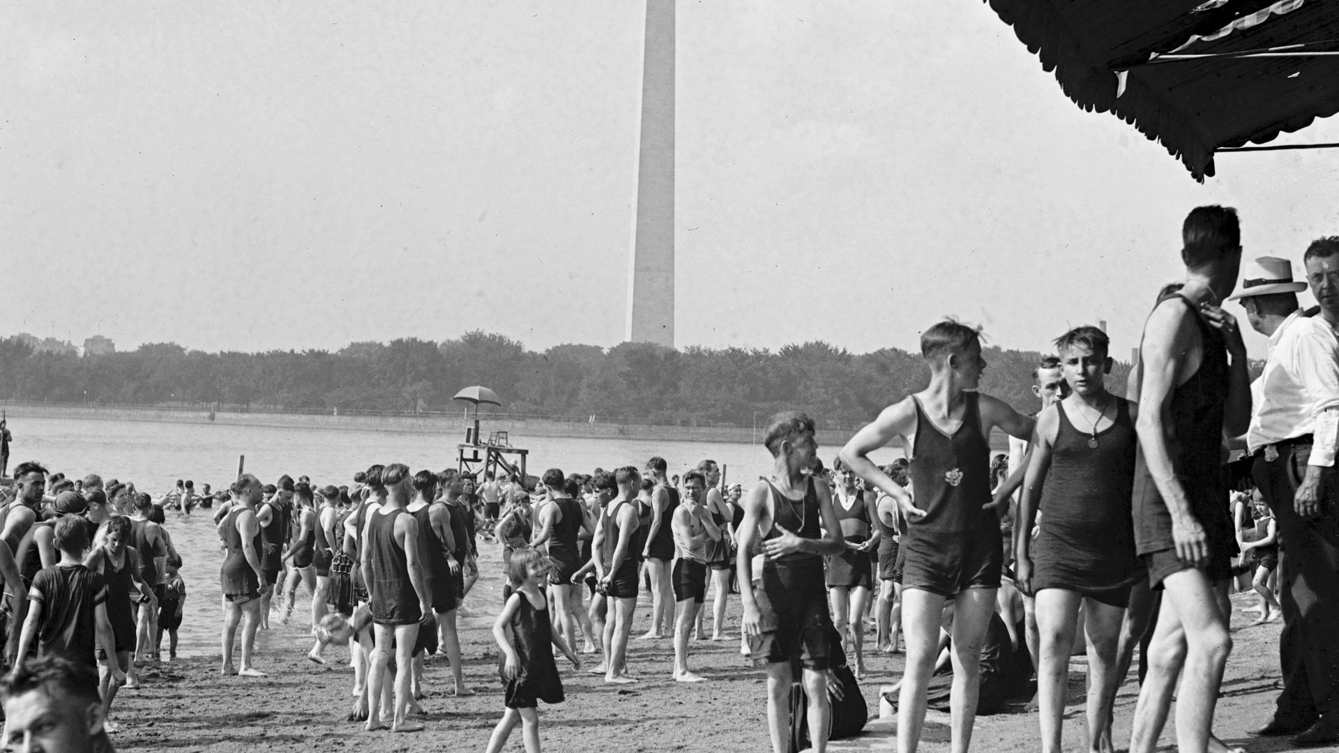 A large crowd of boys in bathing suits on the beach at Washington, DC's tidal basin, in the early twentieth century. The Washington Monument is visible in the background. 