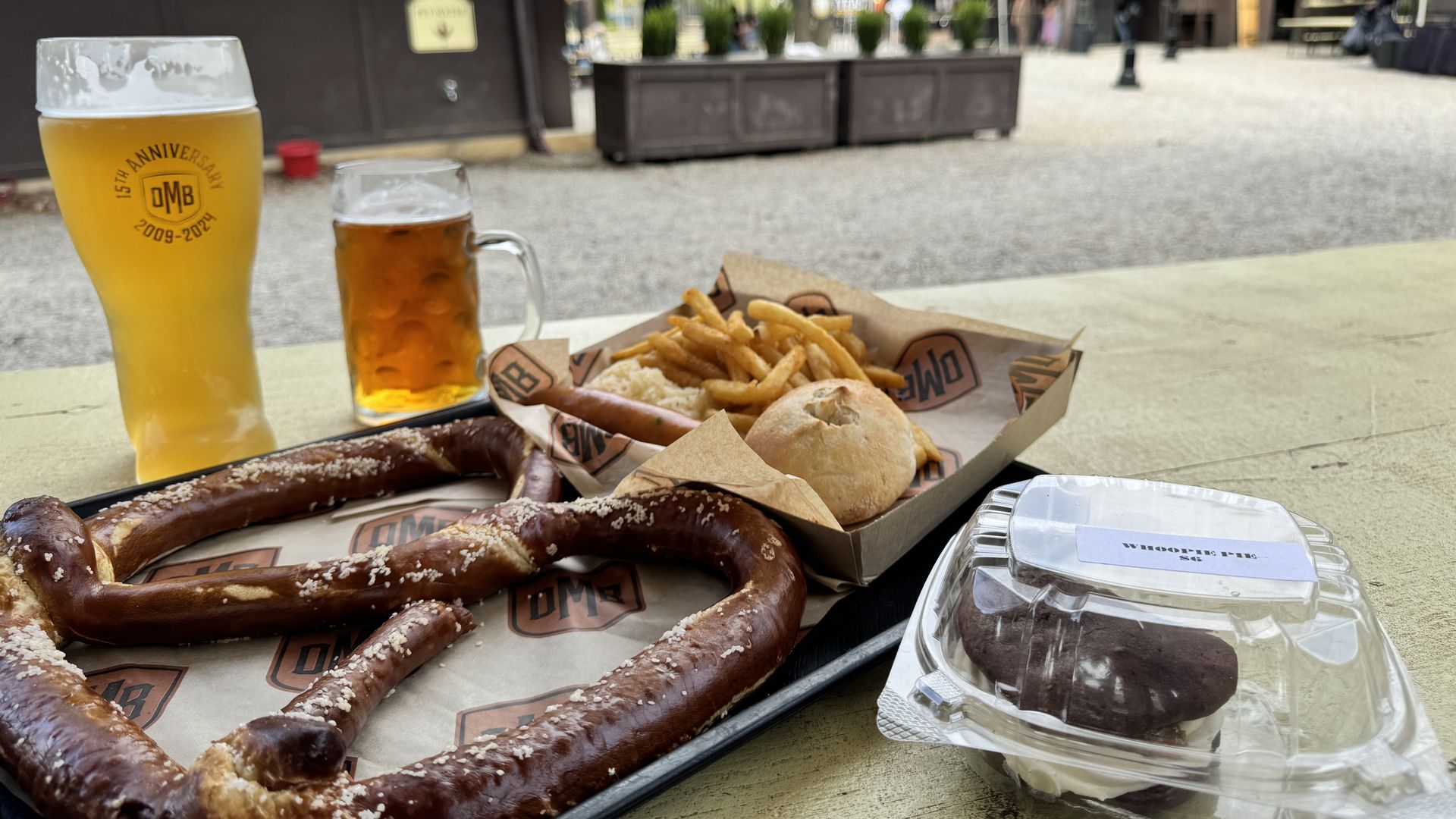 Large soft pretzel with salt, fries and roll in a paper tray, whoopie pie dessert in plastic container, two glasses of beer on an outdoor picnic table in a park setting.