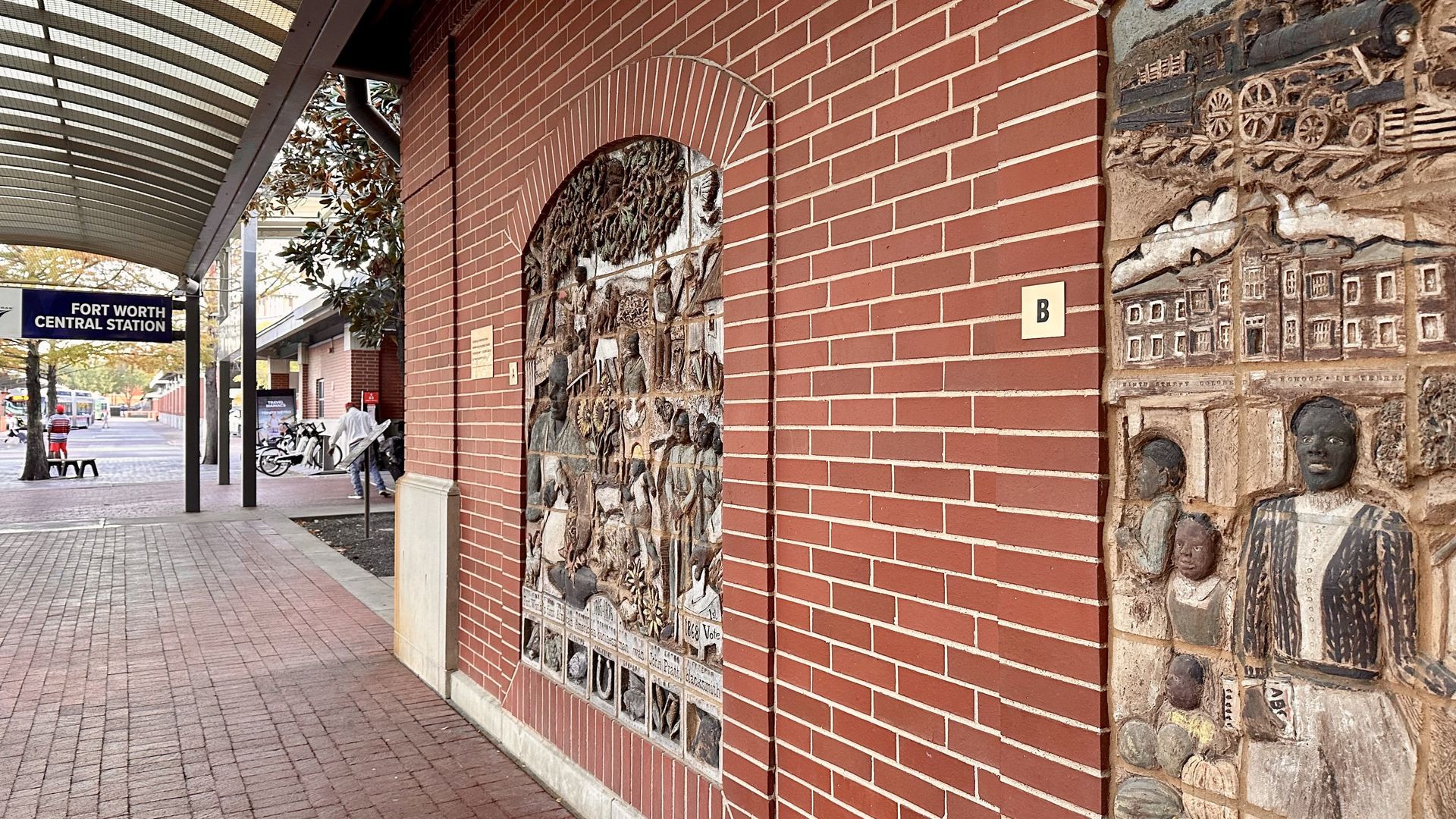 Red brick wall at Fort Worth Central Station with detailed historical bas-relief sculptures depicting people, buildings, and a train. Covered sidewalk and trees visible nearby.
