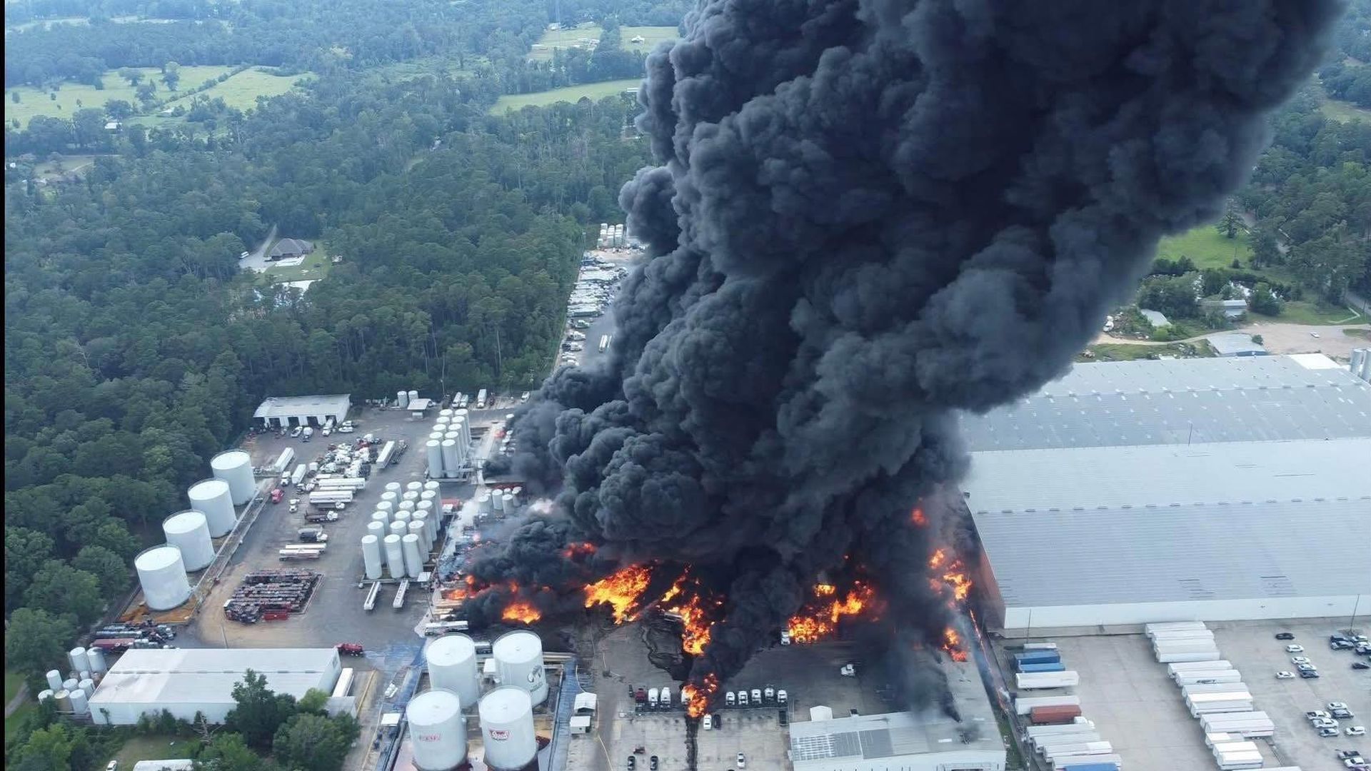 A large fire at an industrial facility with massive black smoke rising over white storage tanks and vehicles, surrounded by green forest and other buildings.