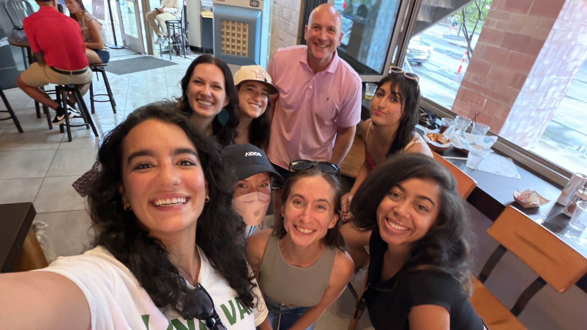 Group of friends taking a cheerful selfie in a casual cafe, with a bald man in a pink shirt center, two women nearby (one wearing a cap) and others leaning in for the photo.