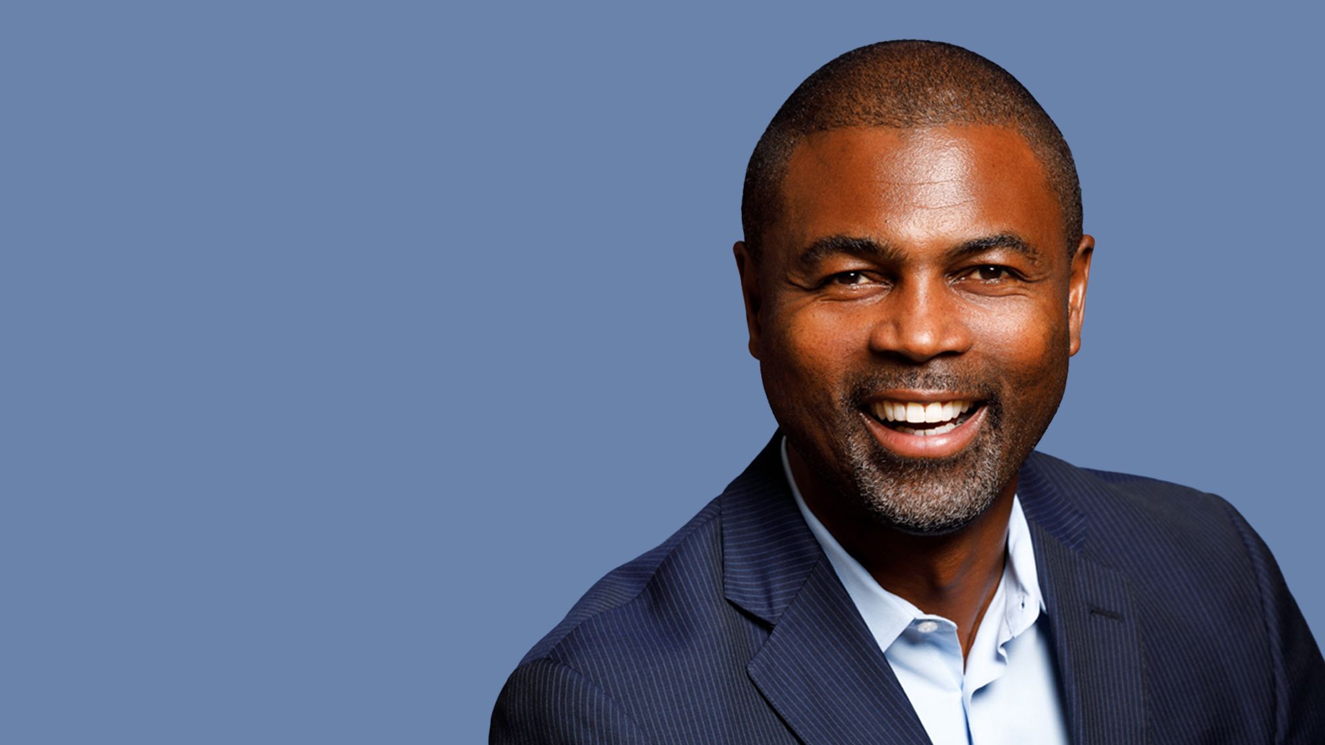 Close-up portrait of a smiling African American man in a navy pinstripe suit and light blue shirt, against a solid blue backdrop.