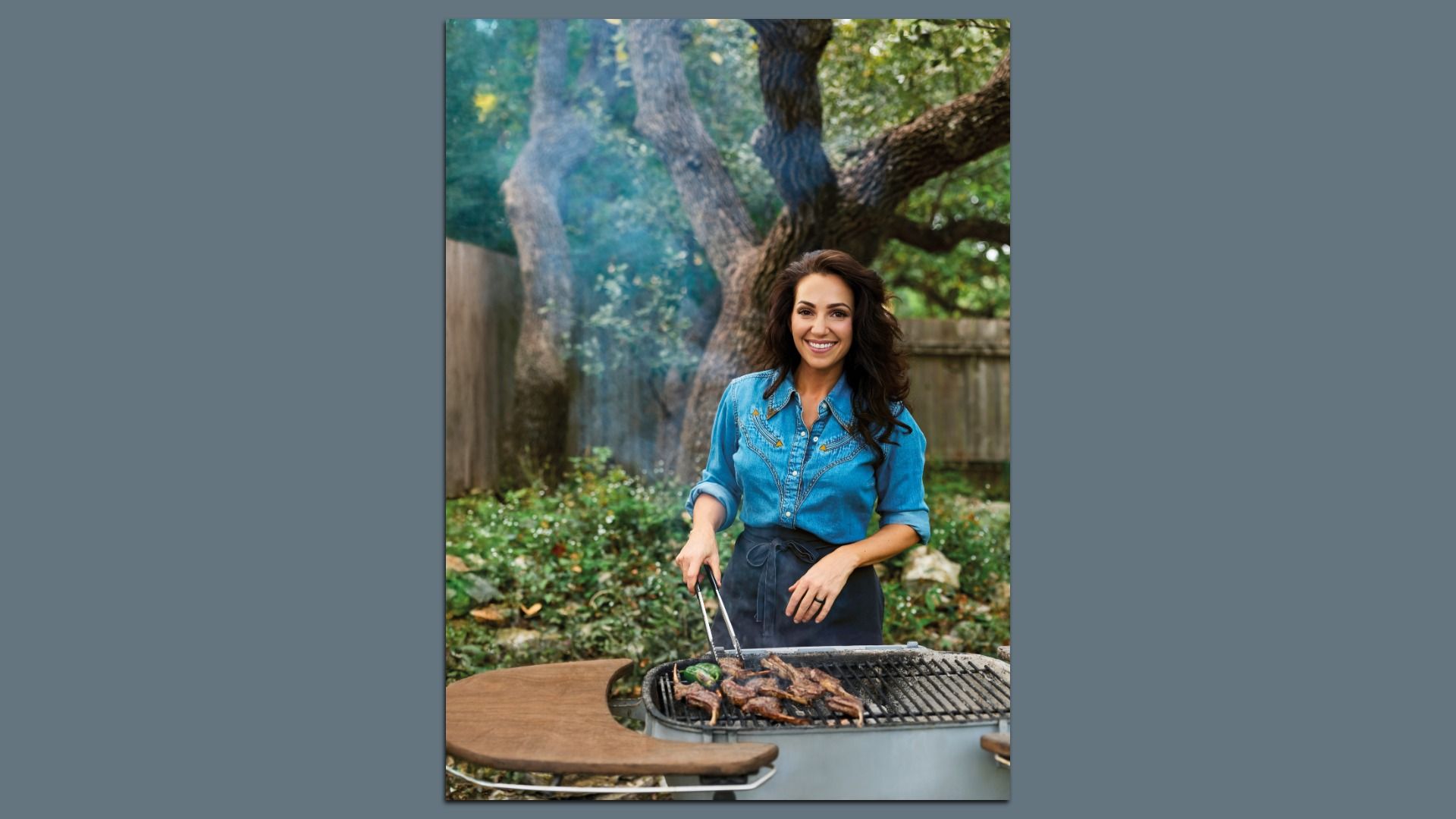 A smiling woman with long dark hair in a blue denim shirt grills meat on an outdoor barbecue, with trees and a wooden fence in a lush garden.