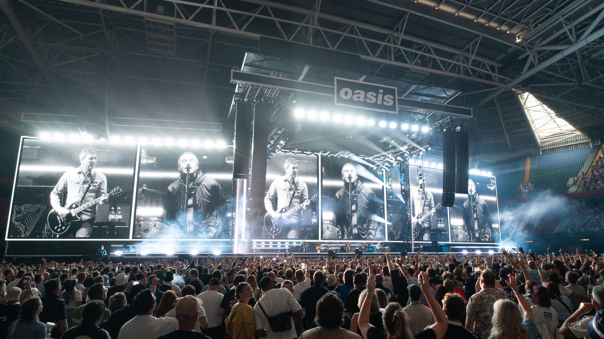 Large crowd at an indoor concert with bright stage lights and big screens showing black-and-white images of musicians performing under a sign reading "oasis".