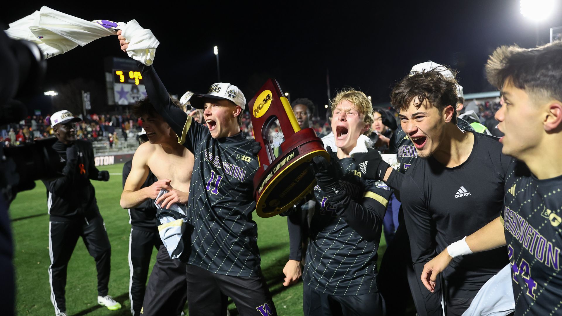 Four to five players celebrate on a soccer pitch, with one waving a t-shirt over his head and another screaming while holding a NCAA trophy. People can be seen in the stands in the background.