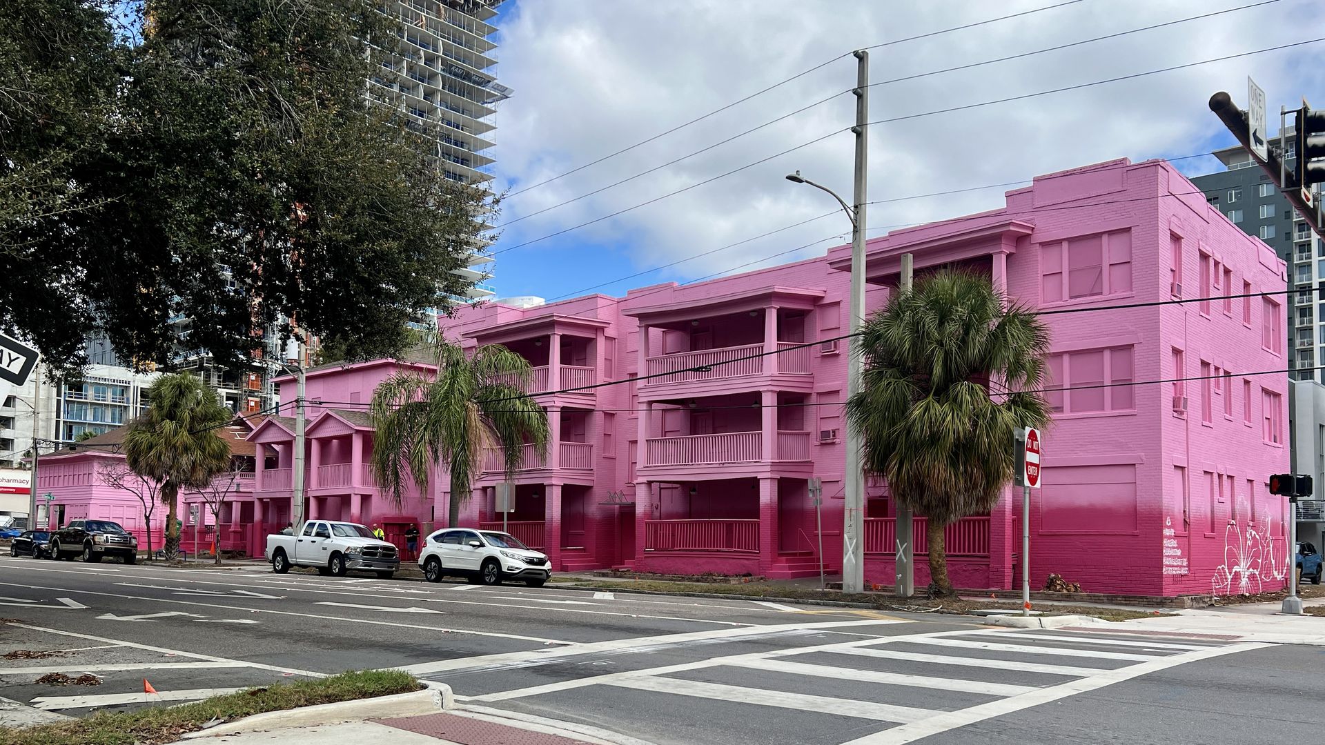City street intersection with a large building painted bright pink, palm trees nearby, several parked cars, and a cloudy blue sky above.