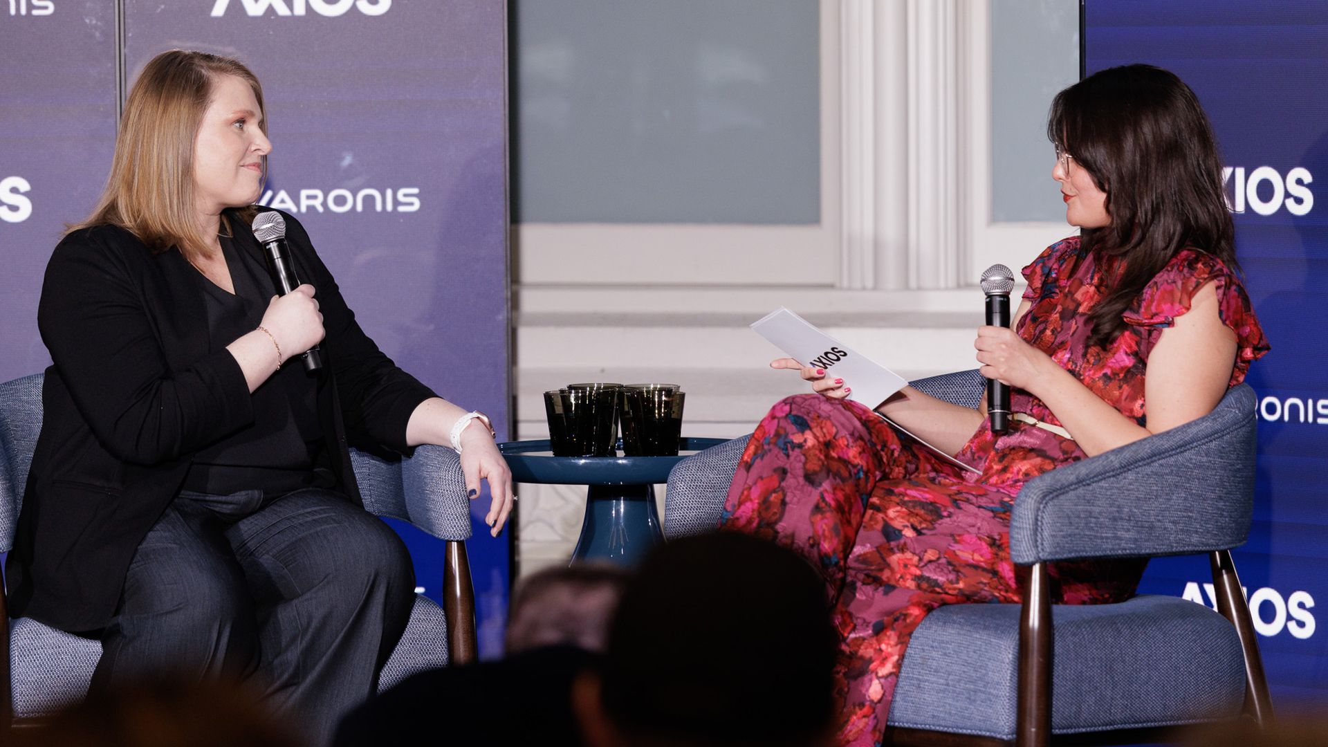 Two women sit on stage in blue chairs, each with a microphone. Left: blonde in black; right: brunette in red floral dress. Small table with three glasses between them; AXIOS and VARONIS logos on backdrop.