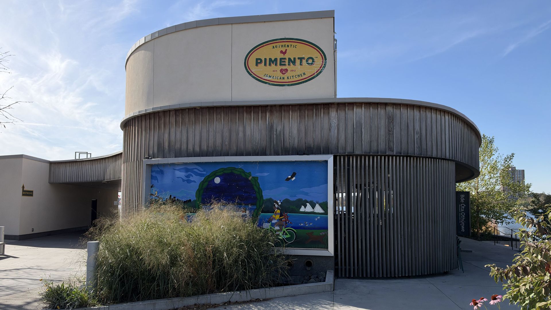Rounded building with a large oval sign reading "PIMENTO" on a pale facade and the phrase "Authentic Jamaican Kitchen." Curved wood siding, clear blue sky, and a mural along the lower wall.