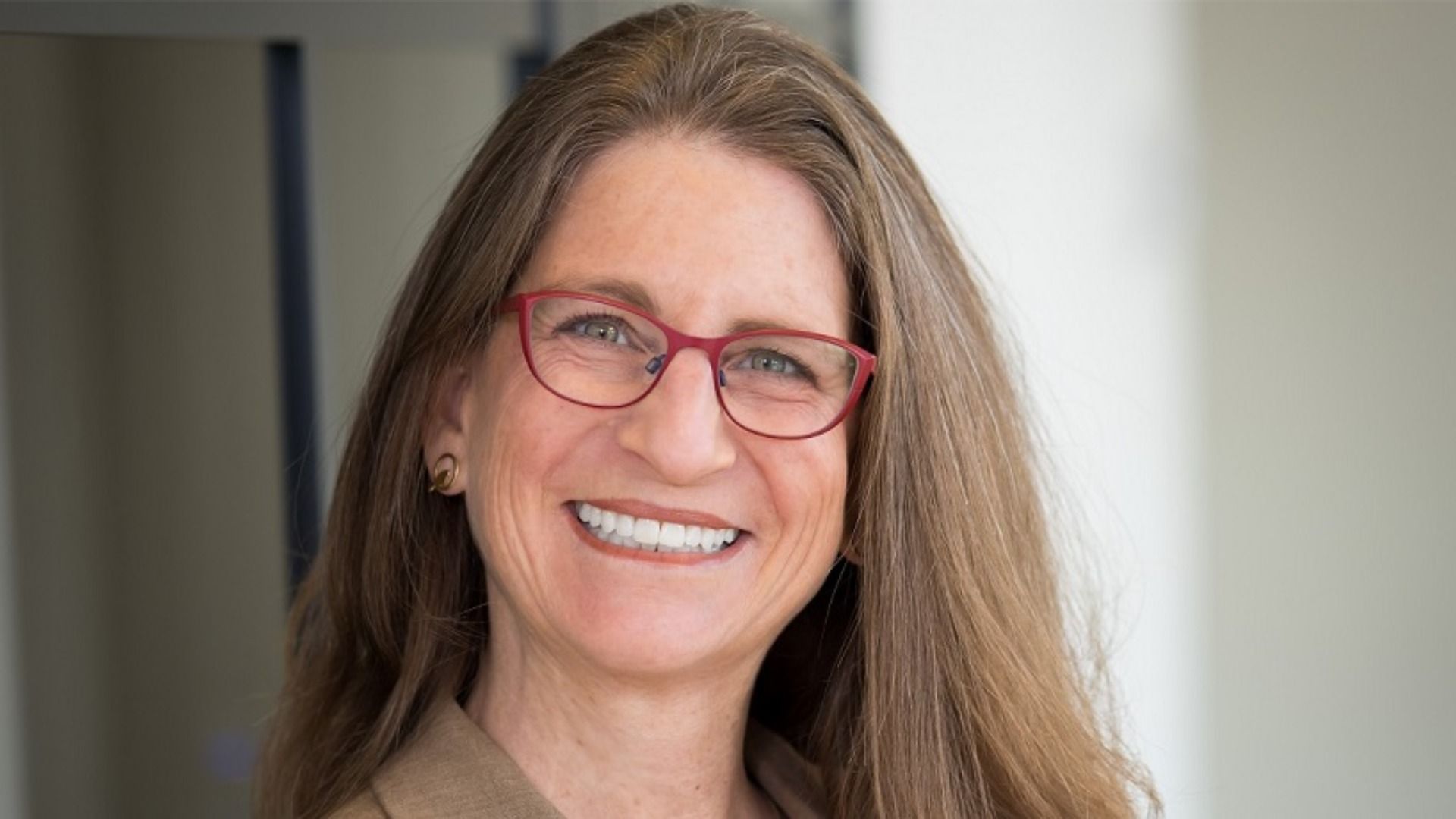 A close-up of a smiling woman with long brown hair, red-framed glasses, and a tan top, against a neutral indoor background.