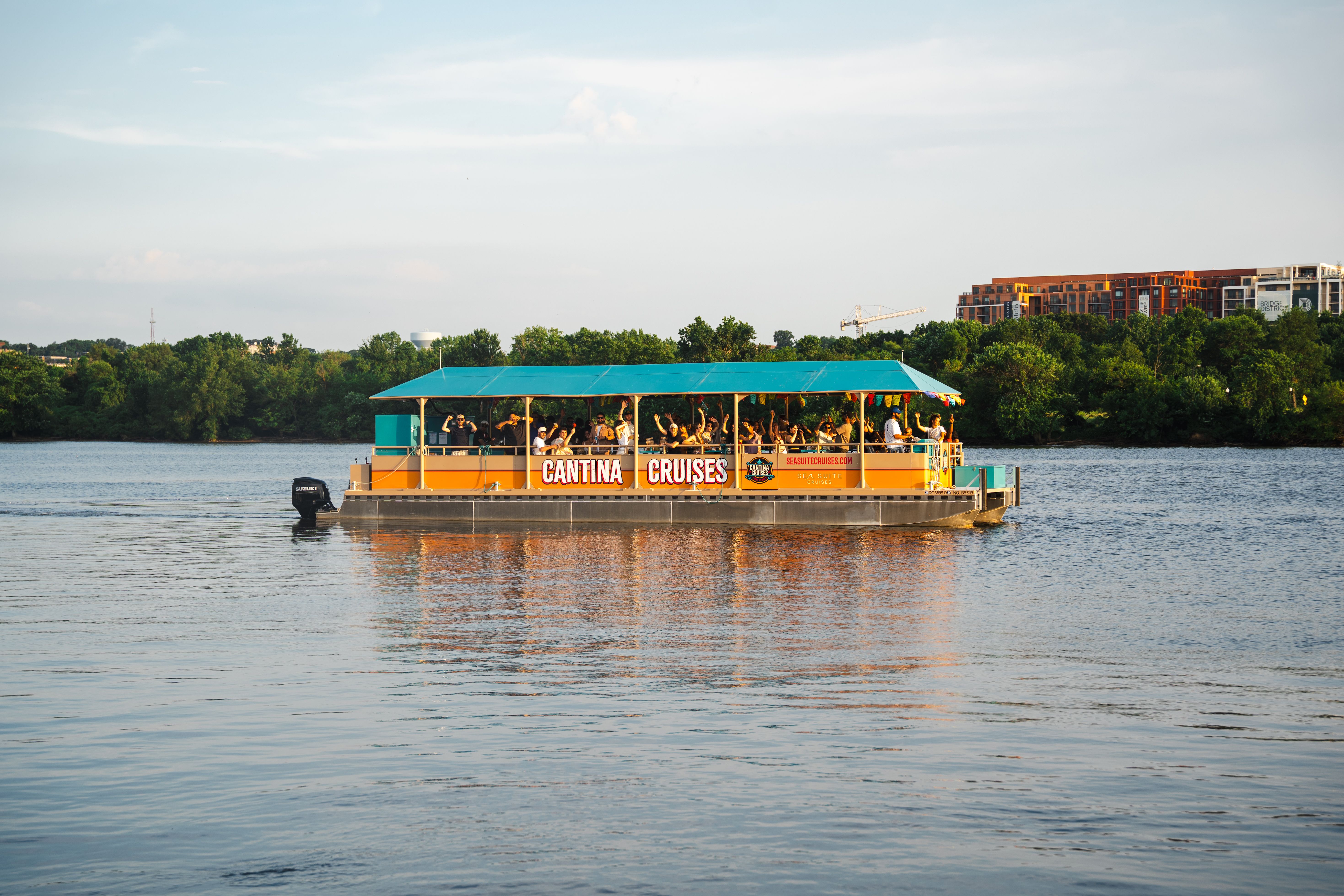 Orange and teal boat labeled Cantina Cruises on a river with many passengers waving, green trees and city buildings in the background under a clear sky.