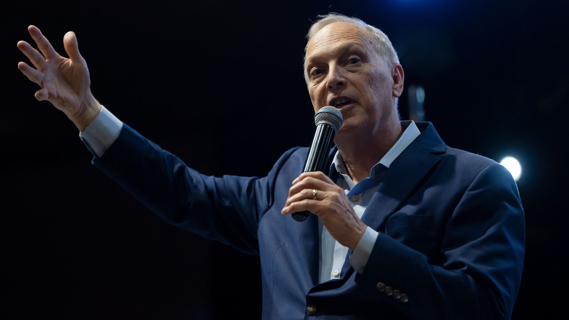 A man in a dark blue suit jacket with a light blue shirt holds a microphone to his mouth with his left hand while holding his right hand up and to his side while he speaks against a dark, black background.