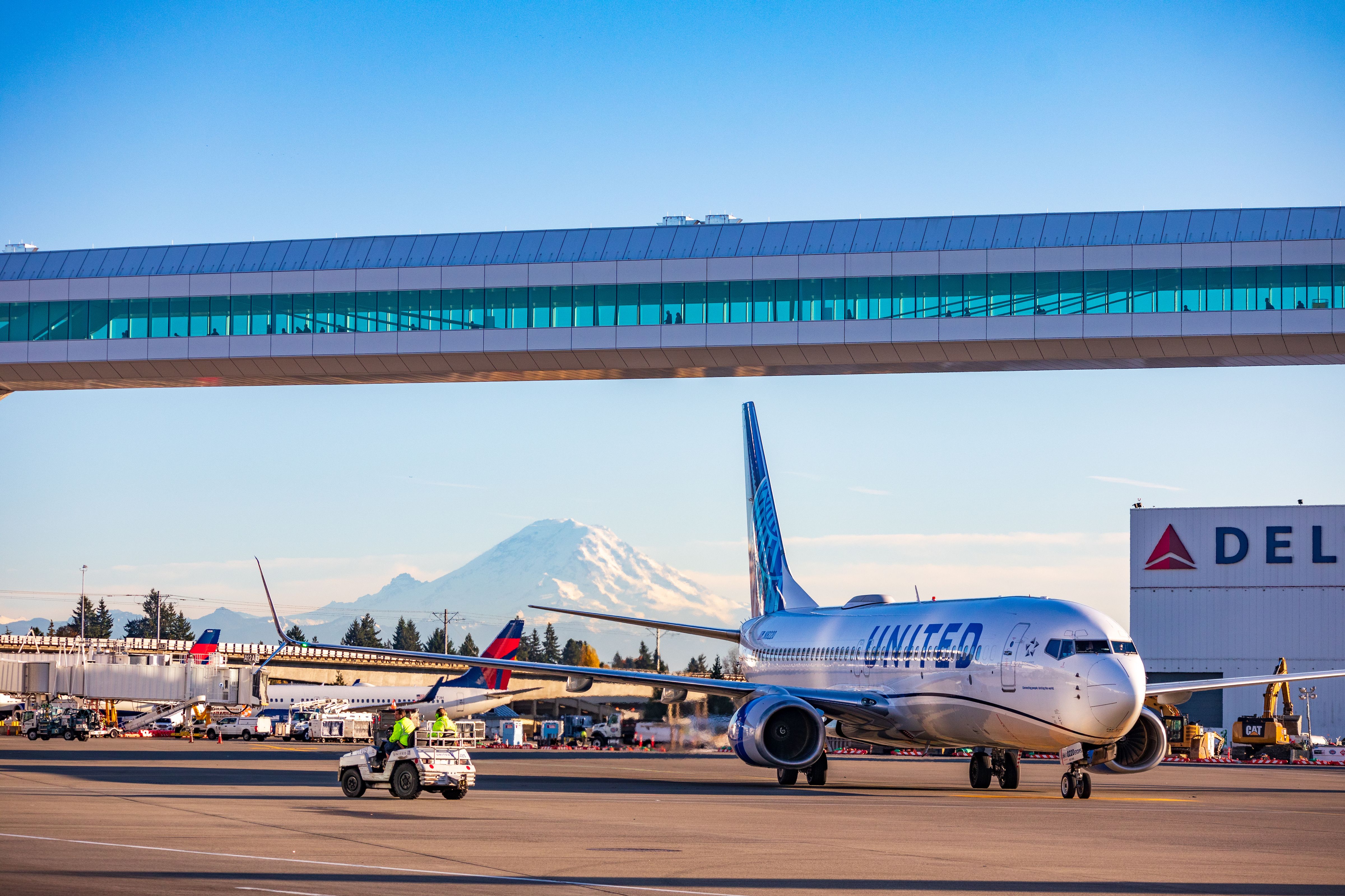 A photo with an airplane, a skywalk and an airport in front of a mountain background. 