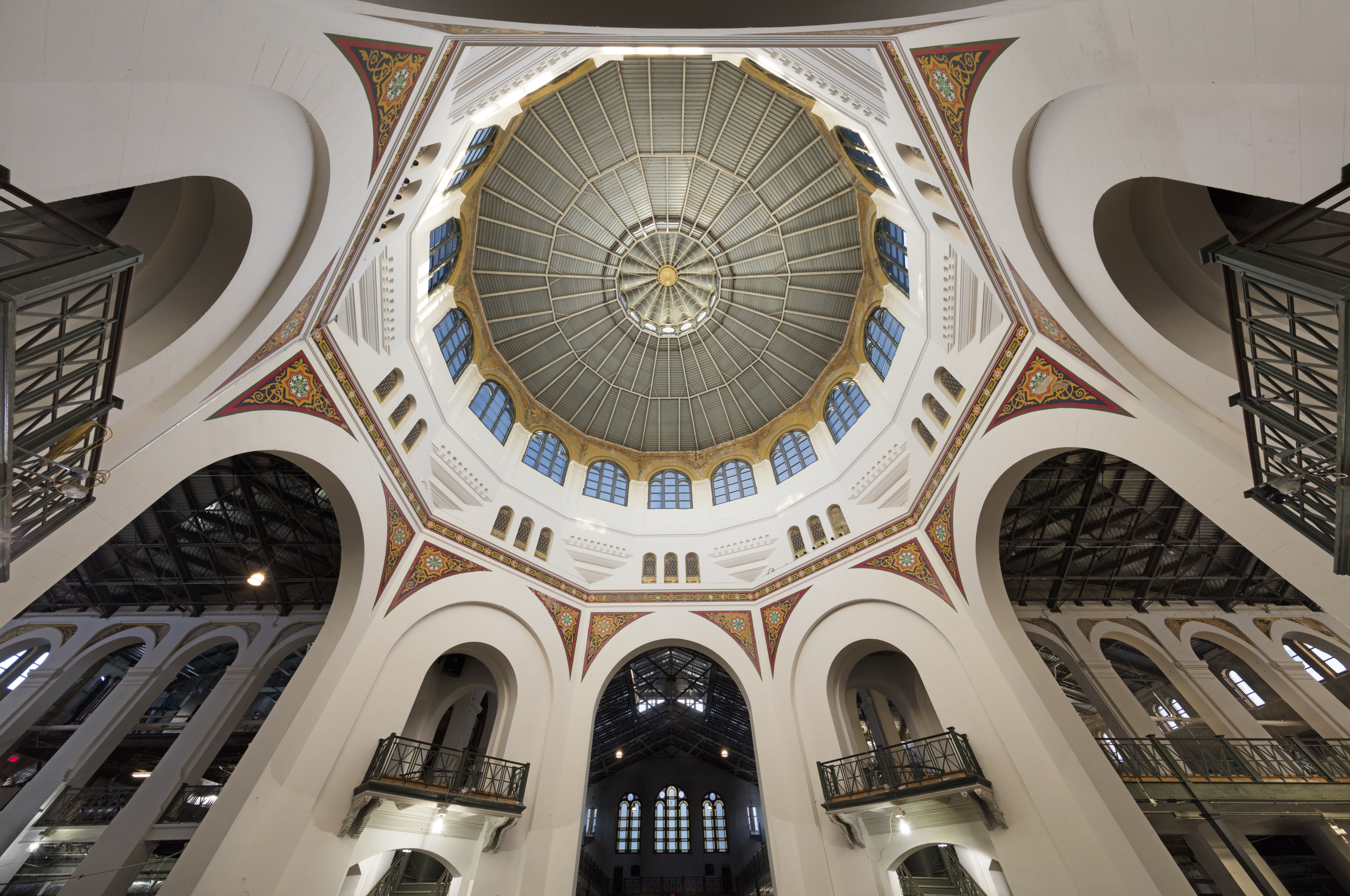 Interior of a grand building with a white octagonal central dome viewed from below; ribbed skylight panels form a circular pattern, surrounded by arched windows and ornate red-and-gold decorations.
