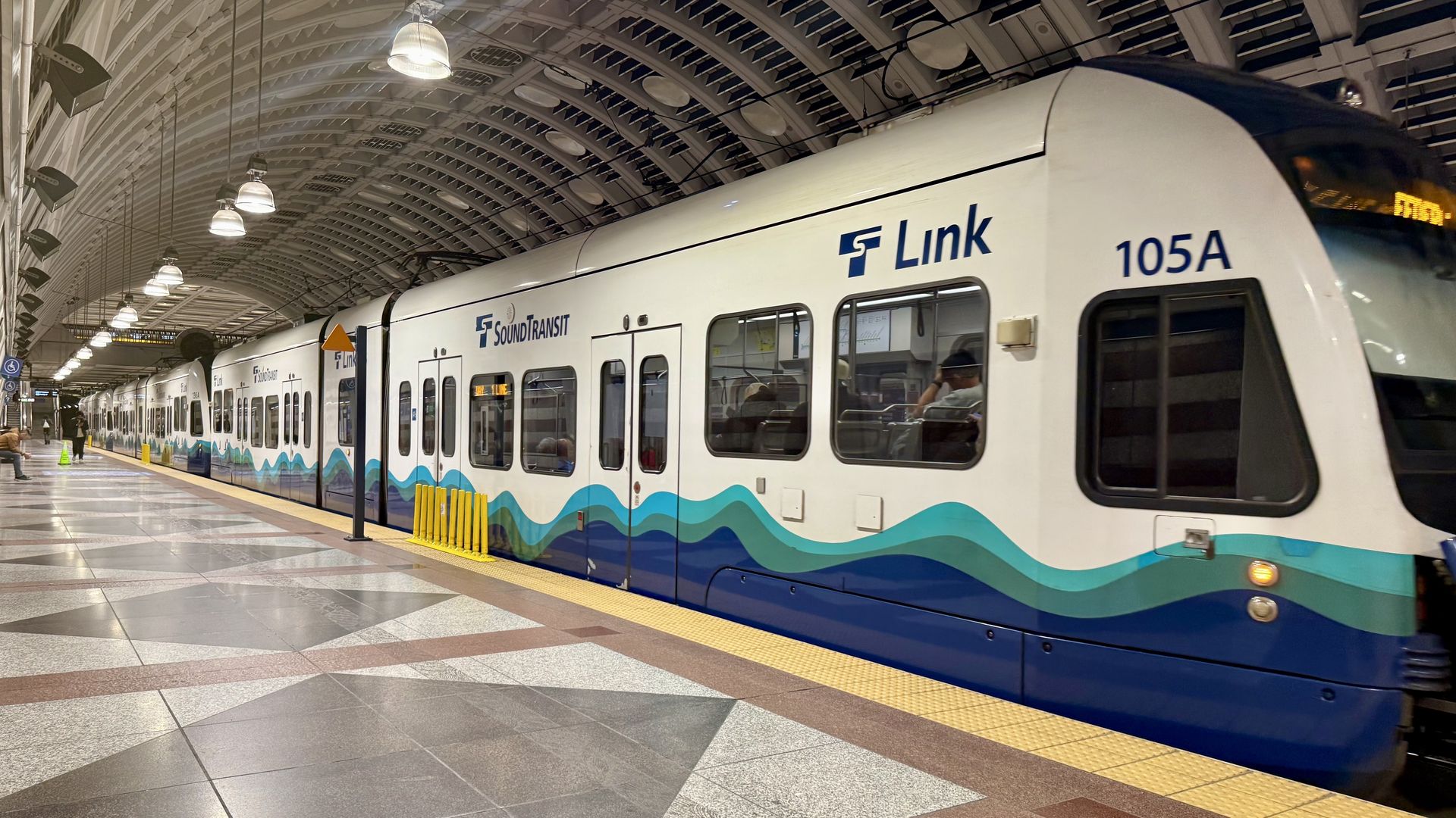 White Link light rail train with blue and turquoise wave graphics at a curved arched-ceiling station. Platform with yellow tactile paving and a few passengers nearby.