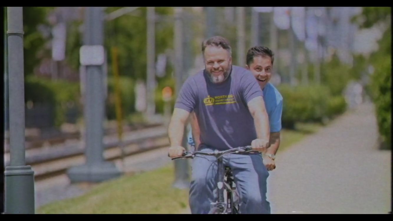 Two men smiling and riding a bicycle together on a sunny day near train tracks and lampposts, with greenery and a sidewalk in the background.