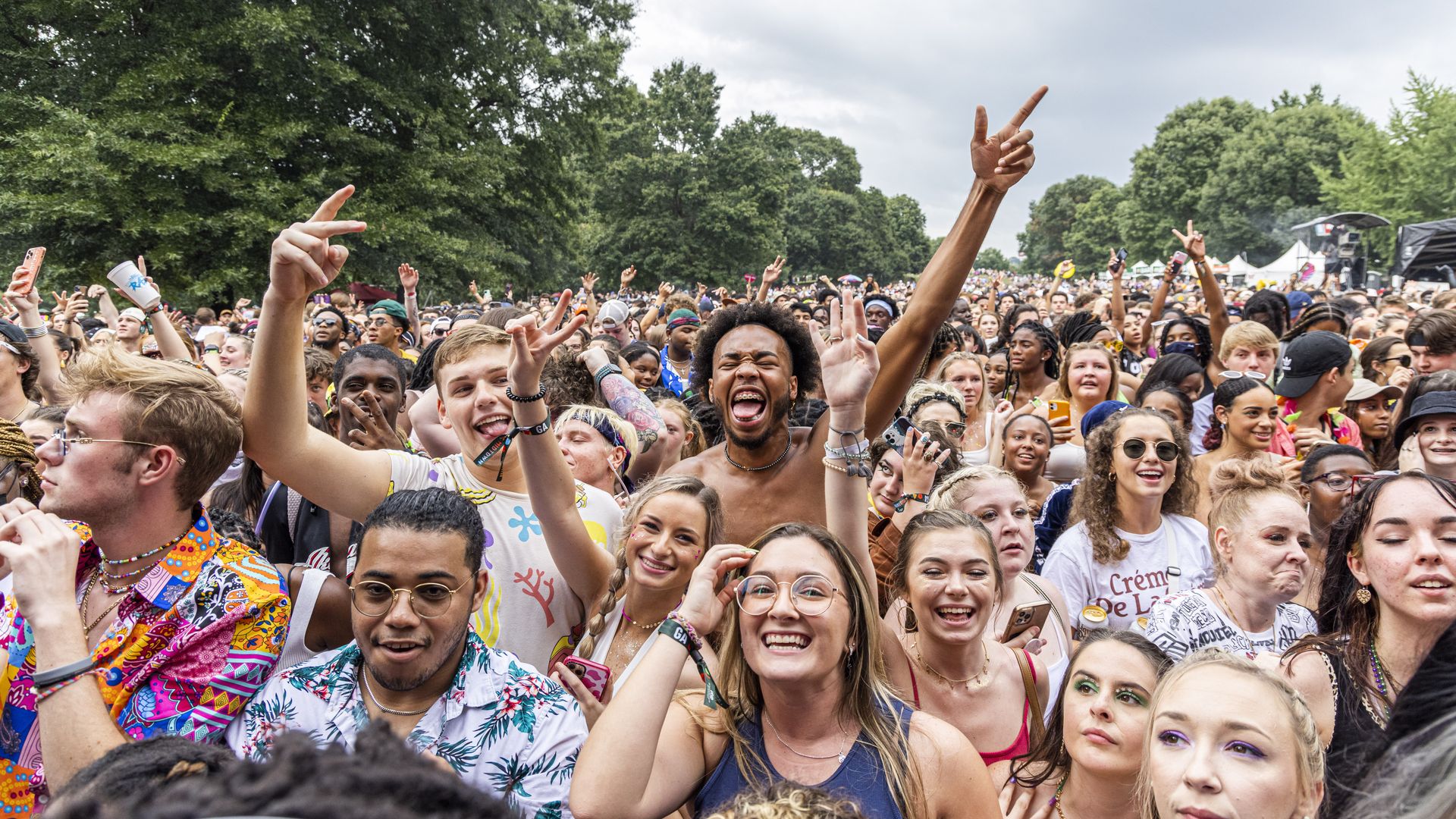 A photograph of a large diverse crowd of mostly young people dancing, smiling and throwing their hands in the air during an outdoor concert