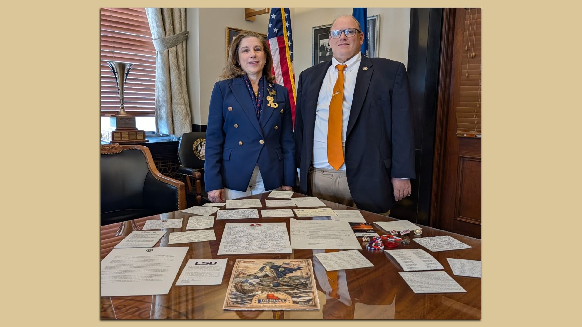 Two people in navy suits stand beside a desk covered with letters in a wood-paneled office, with an American flag and a trophy visible behind them.