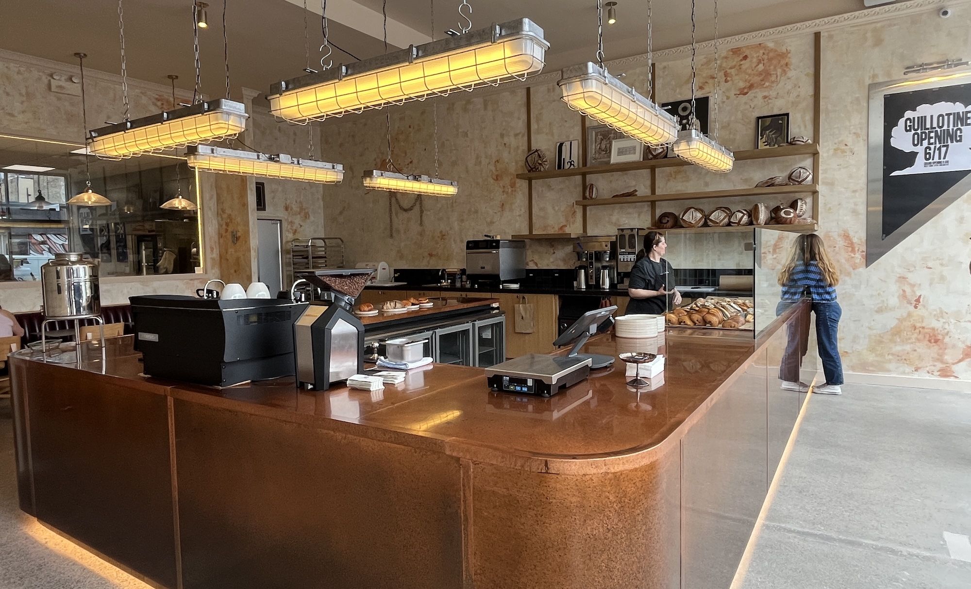 Interior of a cafe bakery with warm yellow hanging lights above a curved brown counter; shelves display bread and pastries as a staff member chats with a customer near the pastry case.