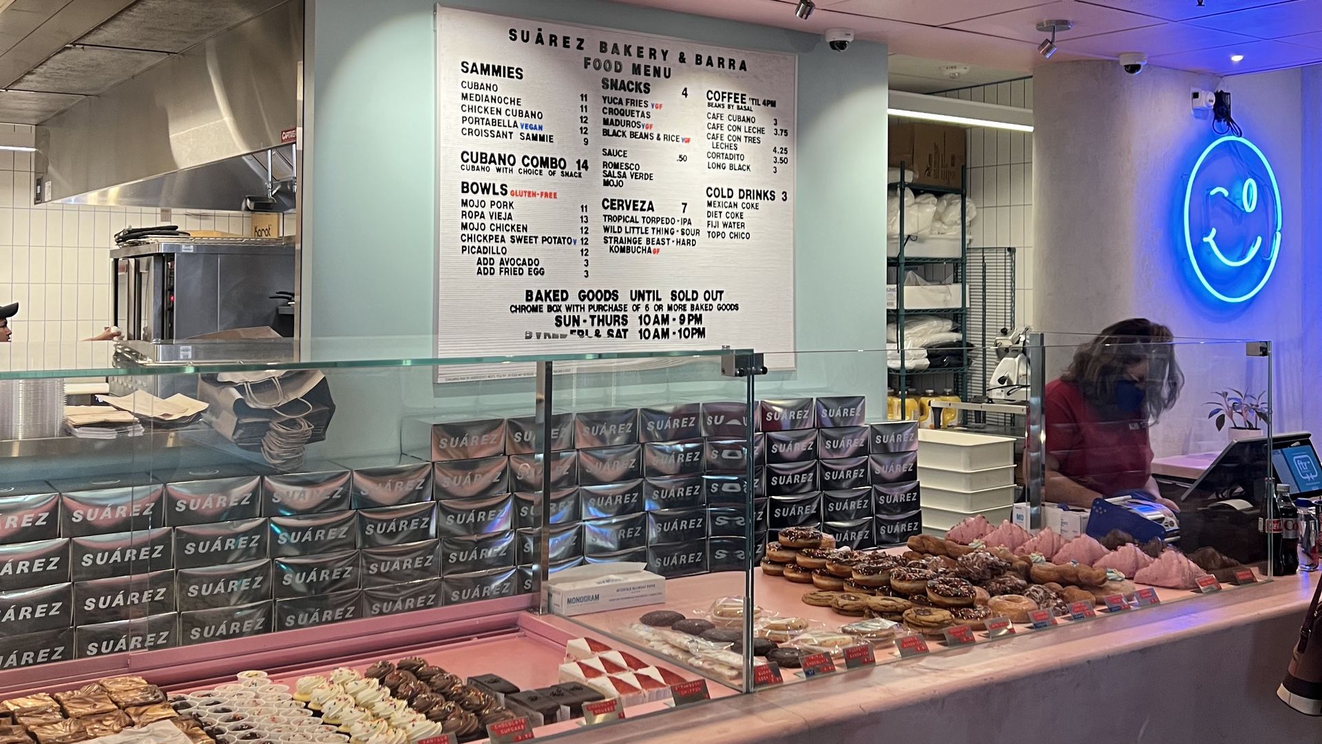 Bakery counter with a pink ceiling, SUÁREZ sign, glass display of donuts and pastries, stacks of SUÁREZ boxes, a menu board, a staff member wearing a mask, and a blue neon smiley sign on the wall.