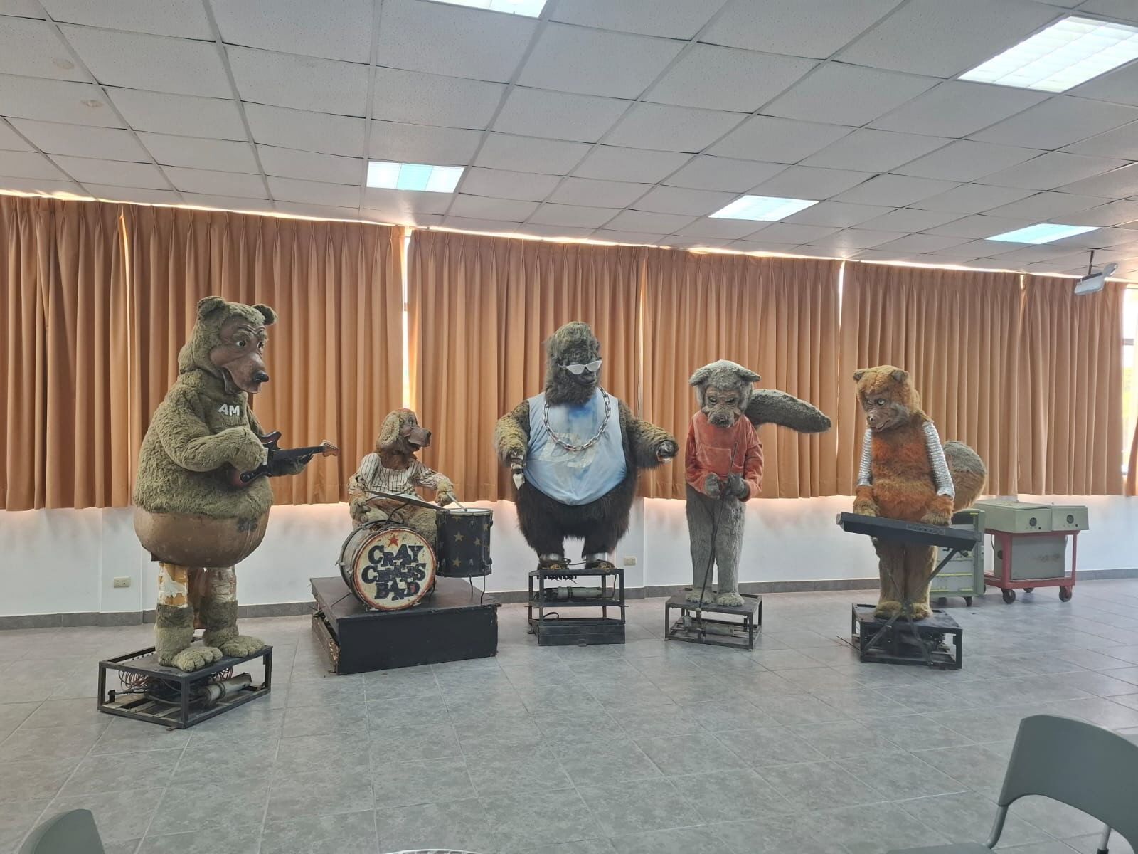 Five animatronic characters perform in a room with brown curtains: left bear with guitar, a drummer, a shaggy center character in sunglasses and chains, a wolf in a red hooded sweatshirt, and a character at a keyboard.