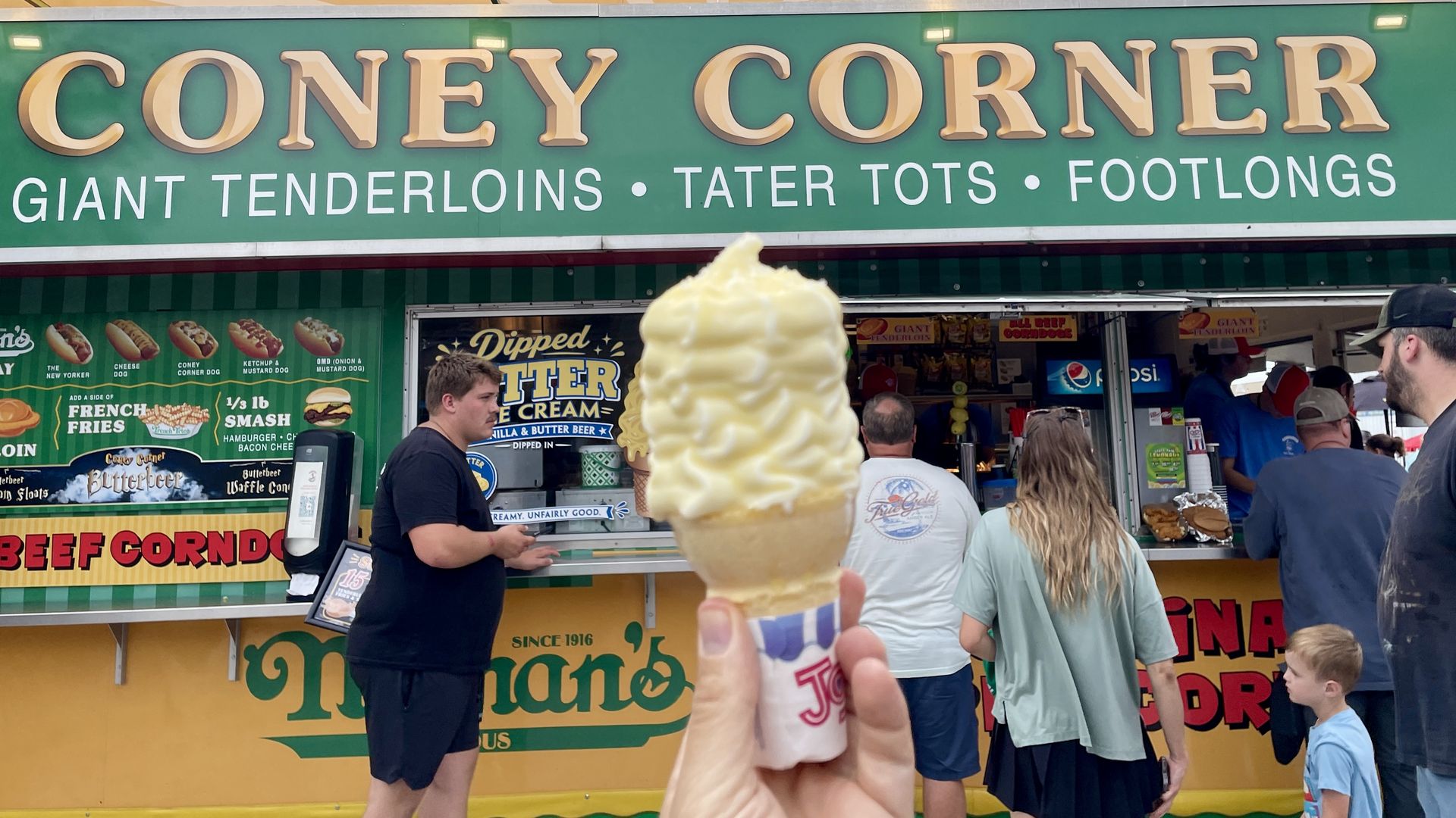 Front view of a green-and-gold Coney Corner stand with large signage; menu includes hot dogs and footlongs. A tall vanilla soft-serve ice cream cone is held up in the foreground as customers order.