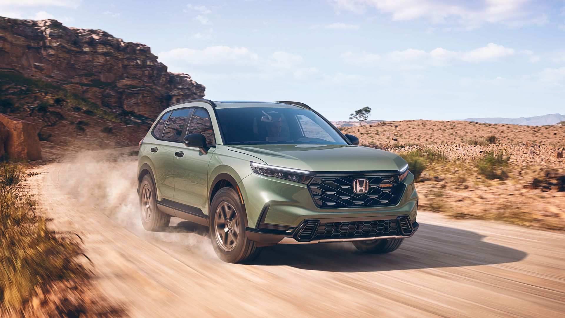 Green Honda CR-V hybrid TrailSport SUV driving on a dusty dirt road in a rocky, desert landscape under a partly cloudy blue sky.