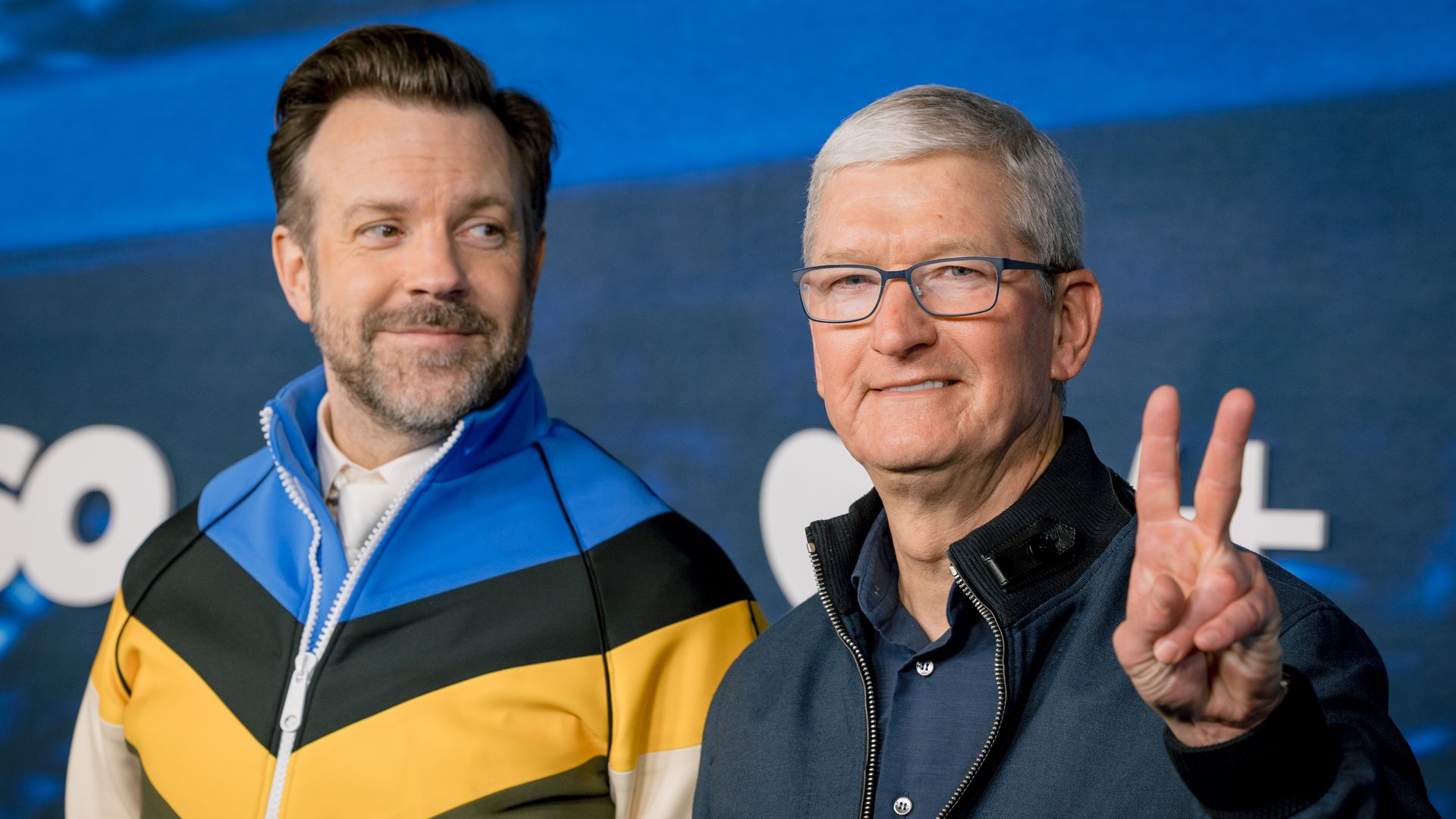 Apple CEO Tim Cook makes a peace sign with his left hand standing next to Jason Sudeilkis