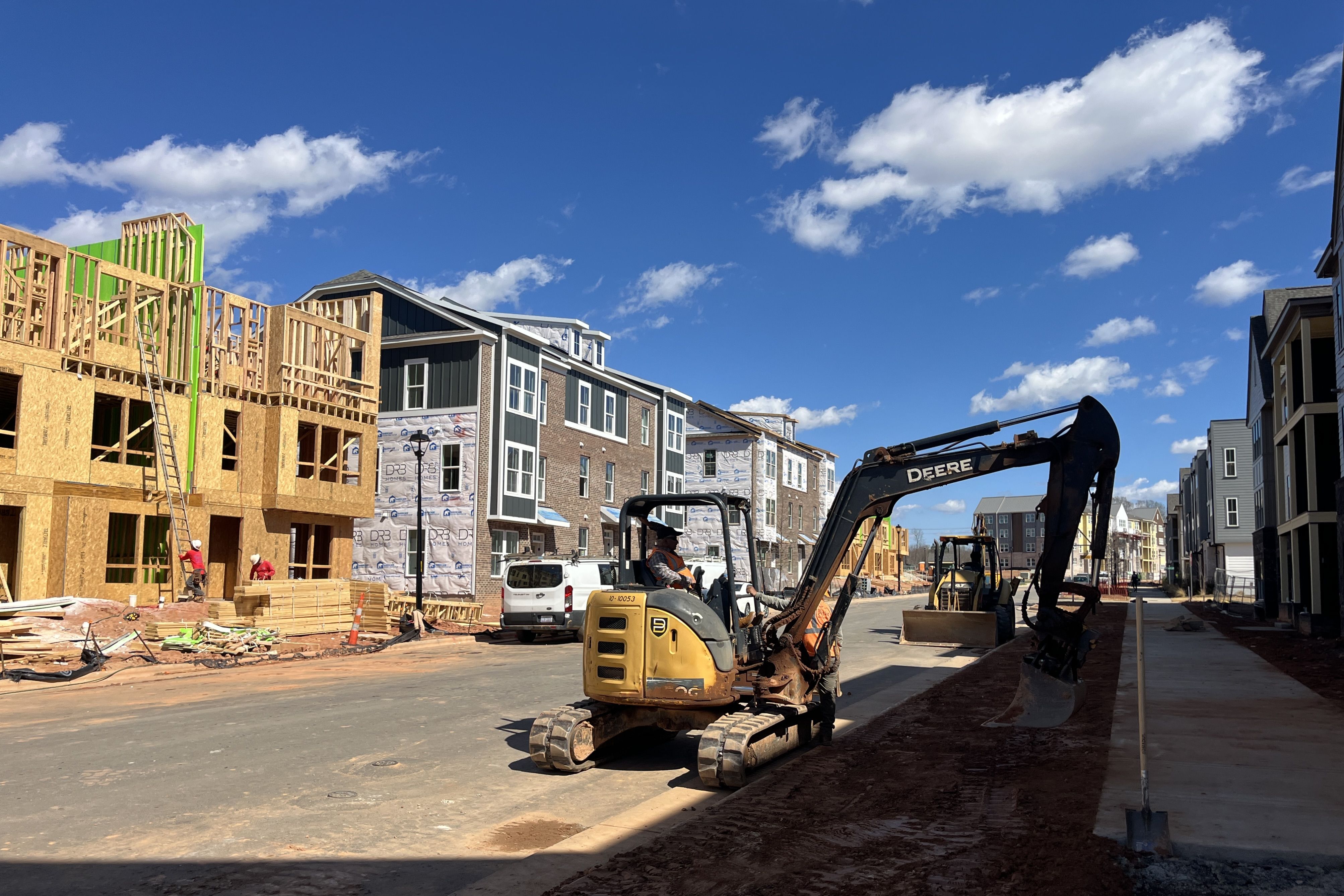 Construction on a residential street: left shows wooden framing for new houses, center rows of modern townhomes, a yellow Deere excavator in the road, workers, and a white van under a bright blue sky.