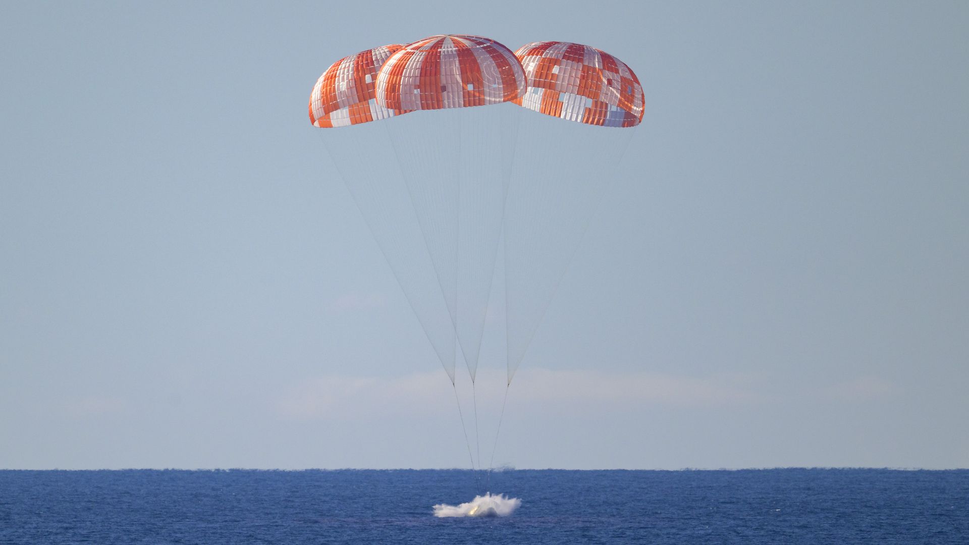 A spacecraft splashes into the deep blue ocean with three orange-and-white parachutes deployed.