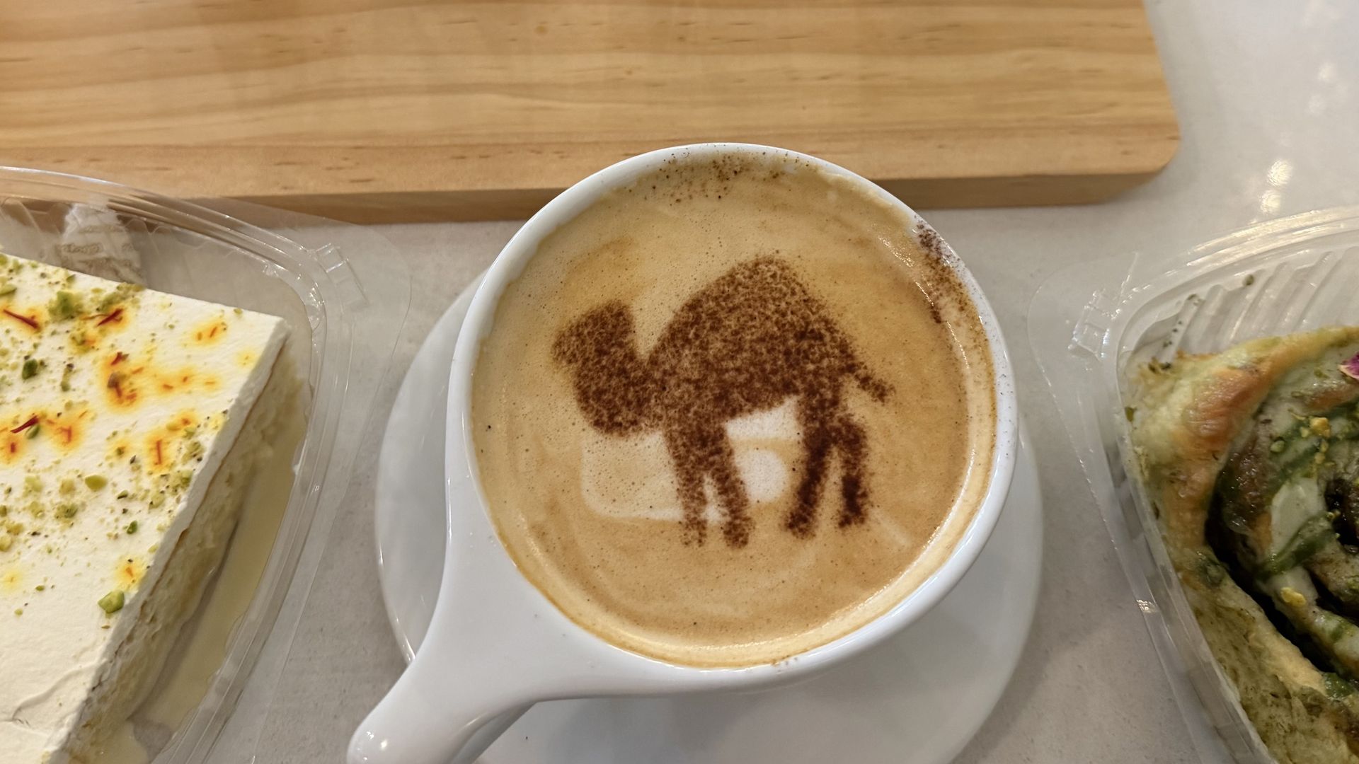 Four glass mugs with varying coffee shades on a wooden tray, a latte with camel-shaped foam art, and two pieces of dessert in plastic containers on a white table.