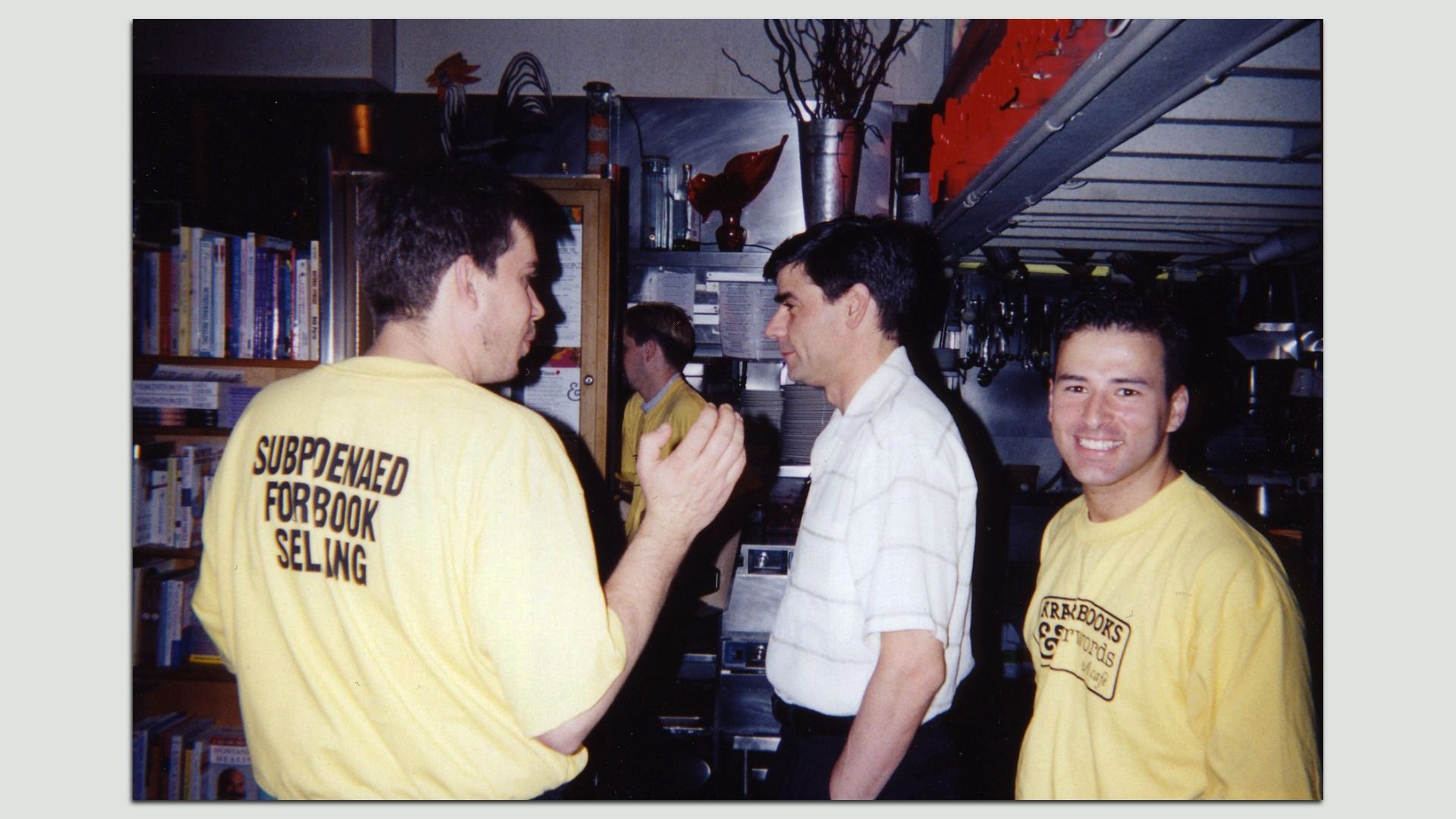 Three men in a busy kitchen. Two wear yellow T-shirts, one with back text "Suspended for Book Selling"; a man in a white shirt faces them. The smiling man on the right looks at the camera.