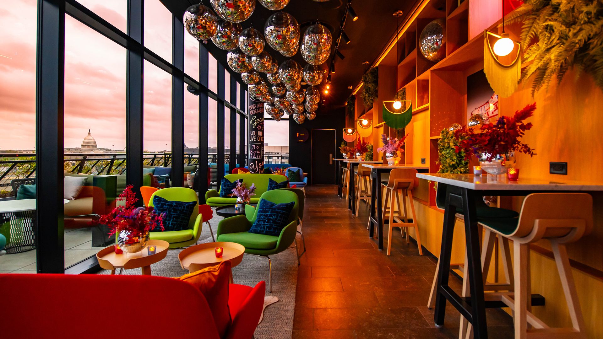 A room with disco balls hanging on the ceiling and colorful lounge chairs overlooking a rooftop with views of the Capitol Building