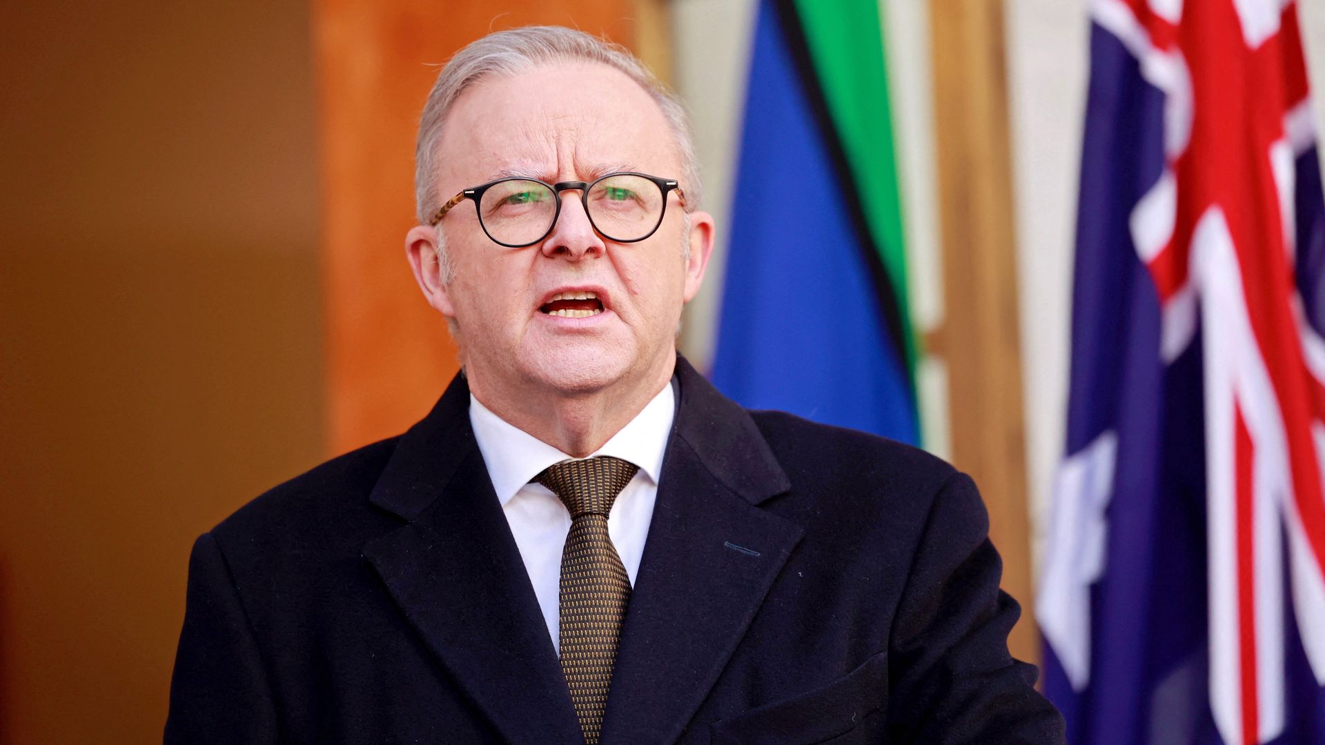 Australia's Prime Minister Anthony Albanese, who has gray hair, glasses, black coat, and patterned tie speaks in front of Australian flags and a green and blue flag. He appears serious during a public address.