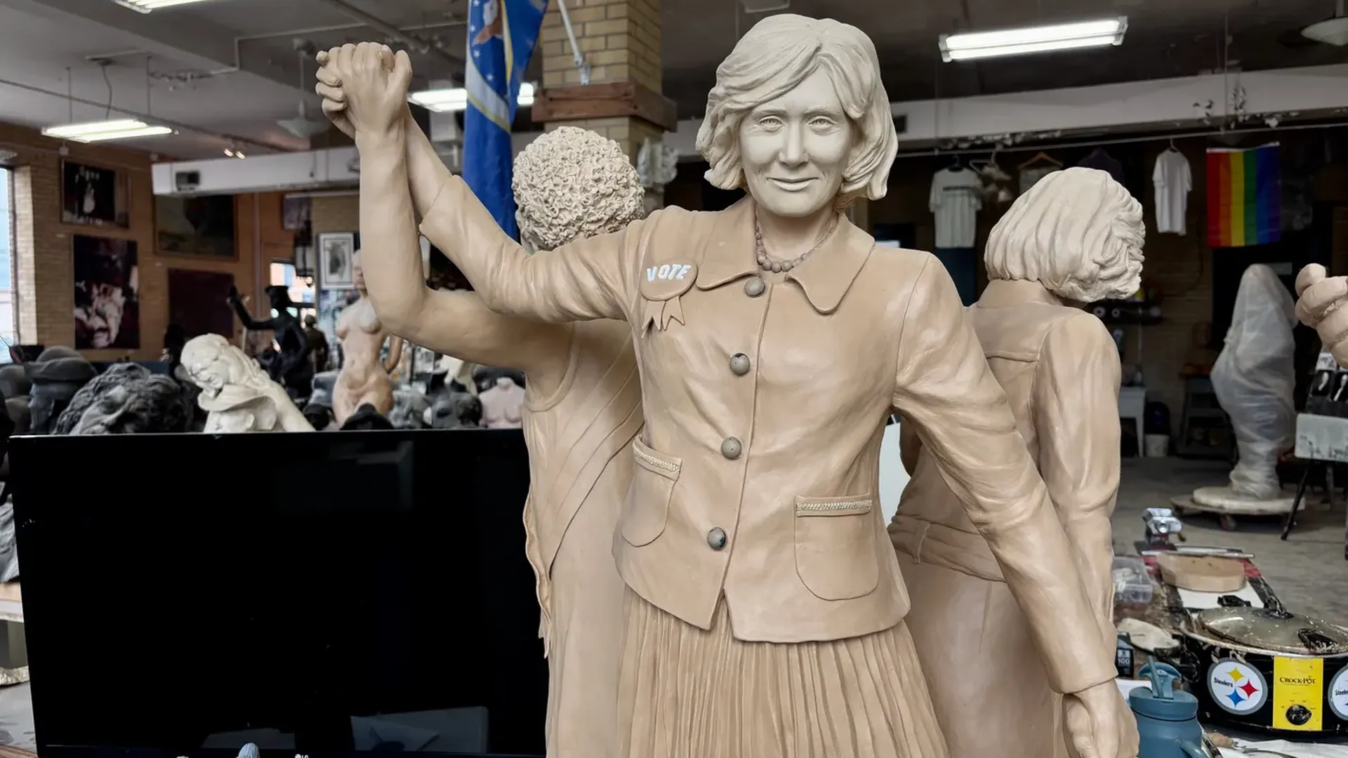 Life-size clay sculpture of a woman in a buttoned coat with a VOTE badge, raising her linked hands with another statue in a busy art studio; a rainbow flag hangs in the background.