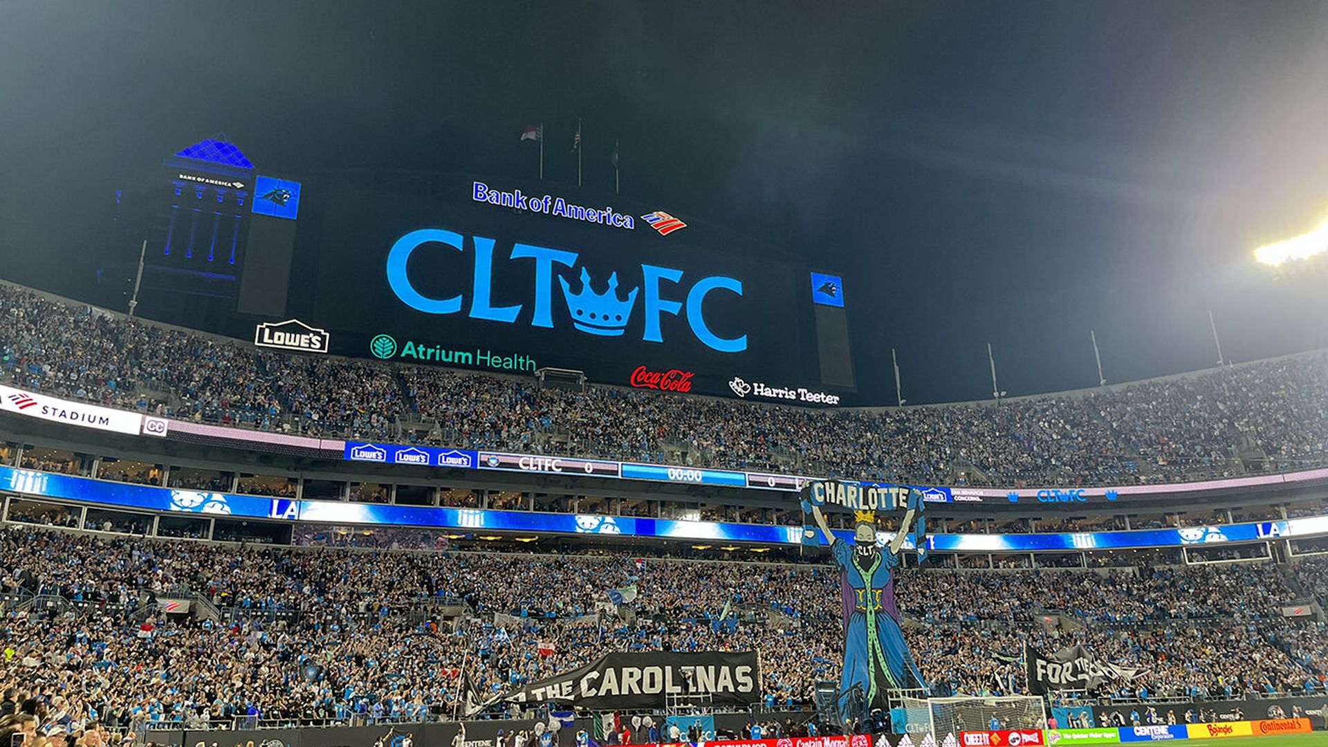 Large soccer stadium filled with fans at night, displaying a giant blue sign "CLT FC" with sponsor logos. Fans hold banners and a large figure with "Charlotte" on scarf.