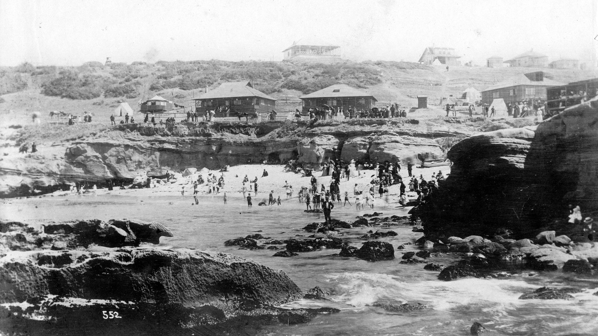 Black-and-white panoramic beach scene: rocky foreground, a large crowd of early 20th-century swimmers and walkers, with wooden beach cottages on grassy dunes in the distance.