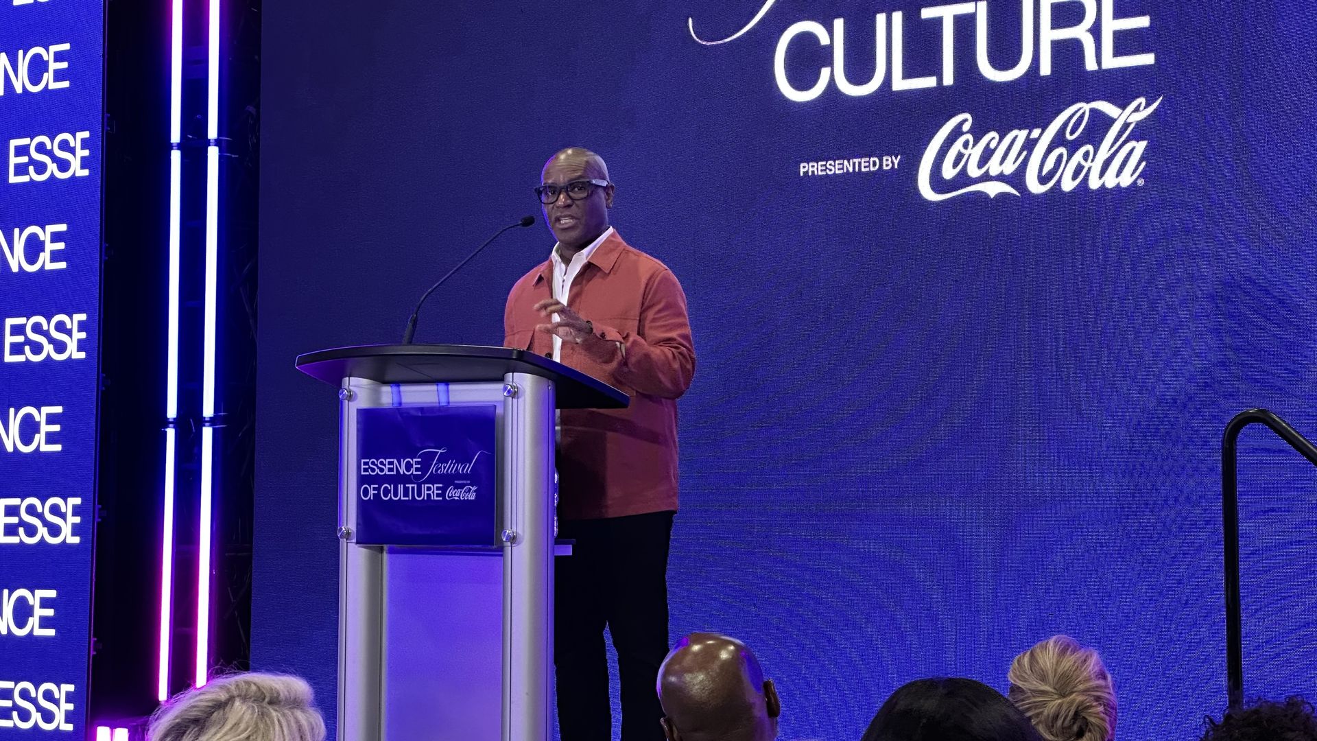 A man in a red jacket and glasses speaks at a podium on a blue stage with a large sign reading "CULTURE"; left banners show "ESSENCE" and "PRESENTED BY Coca-Cola".