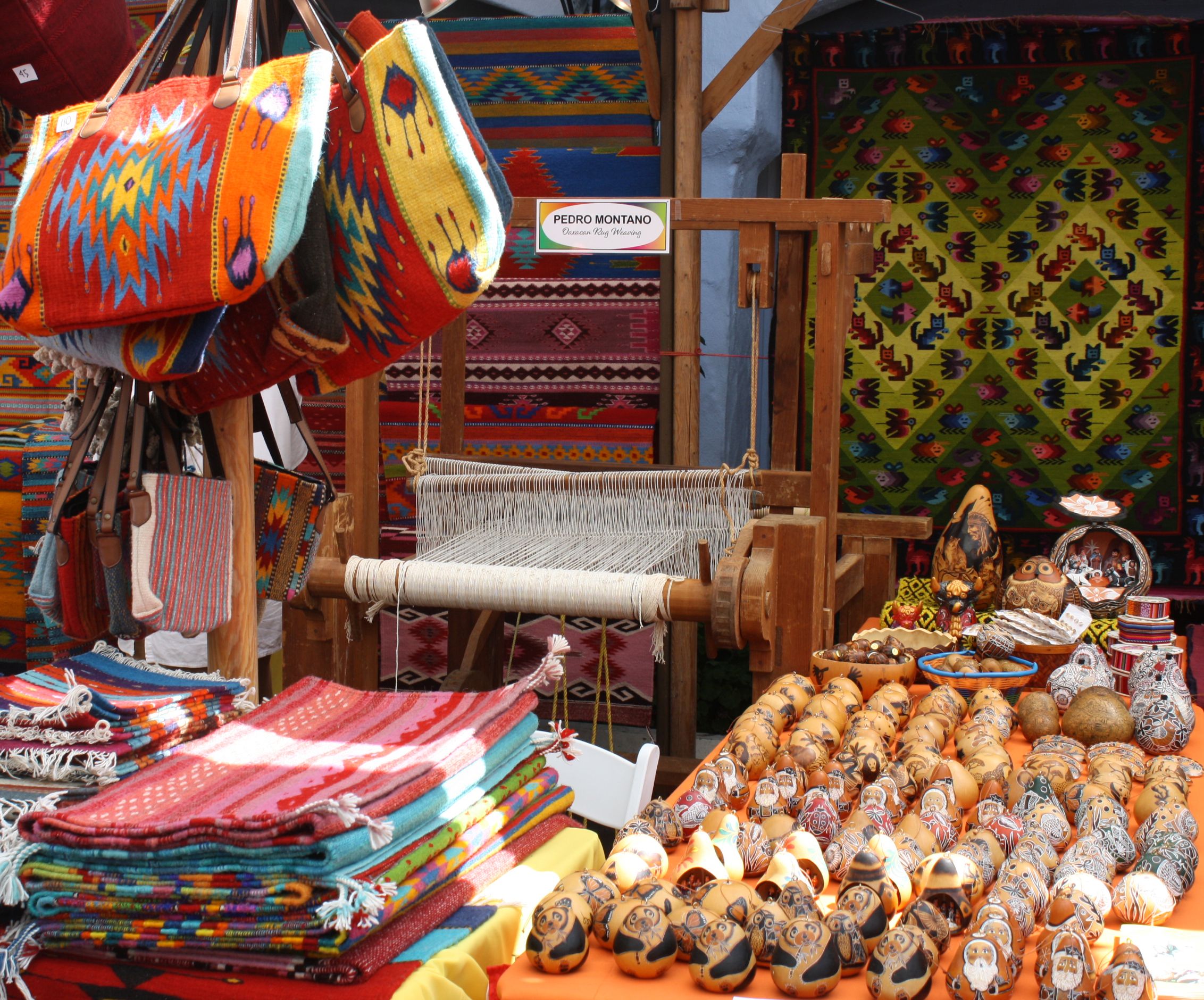 Colorful woven textiles, bags, and blankets displayed at a market with a wooden loom labeled "Pedro Montano Oaxaca Rug Weaving" and decorated gourds on a table.
