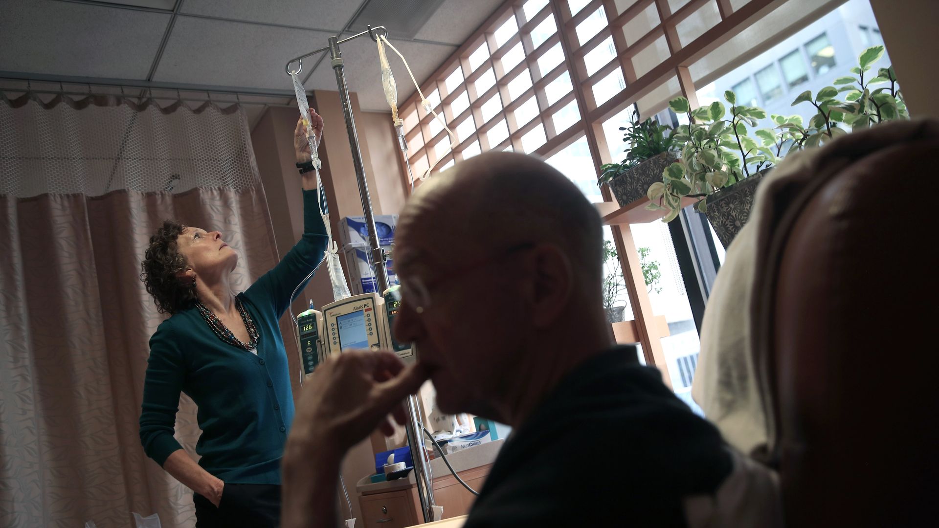 A man in the foreground sits in a chair receiving an IV infusion, and a woman in the background looks at the man's IV pole.