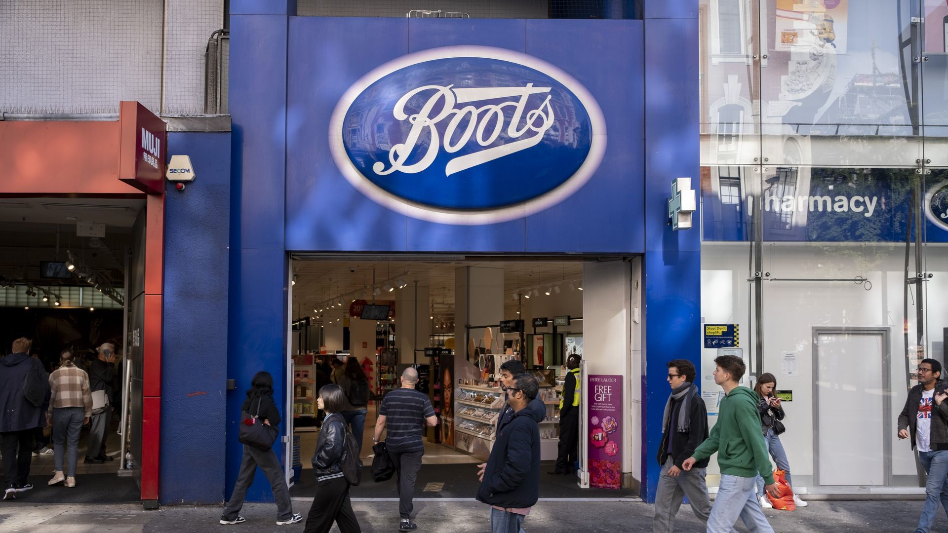 Busy city street with a large Boots storefront. A blue oval Boots sign dominates the center; glass storefront on the right shows a pharmacy sign. People walk past the entrance.