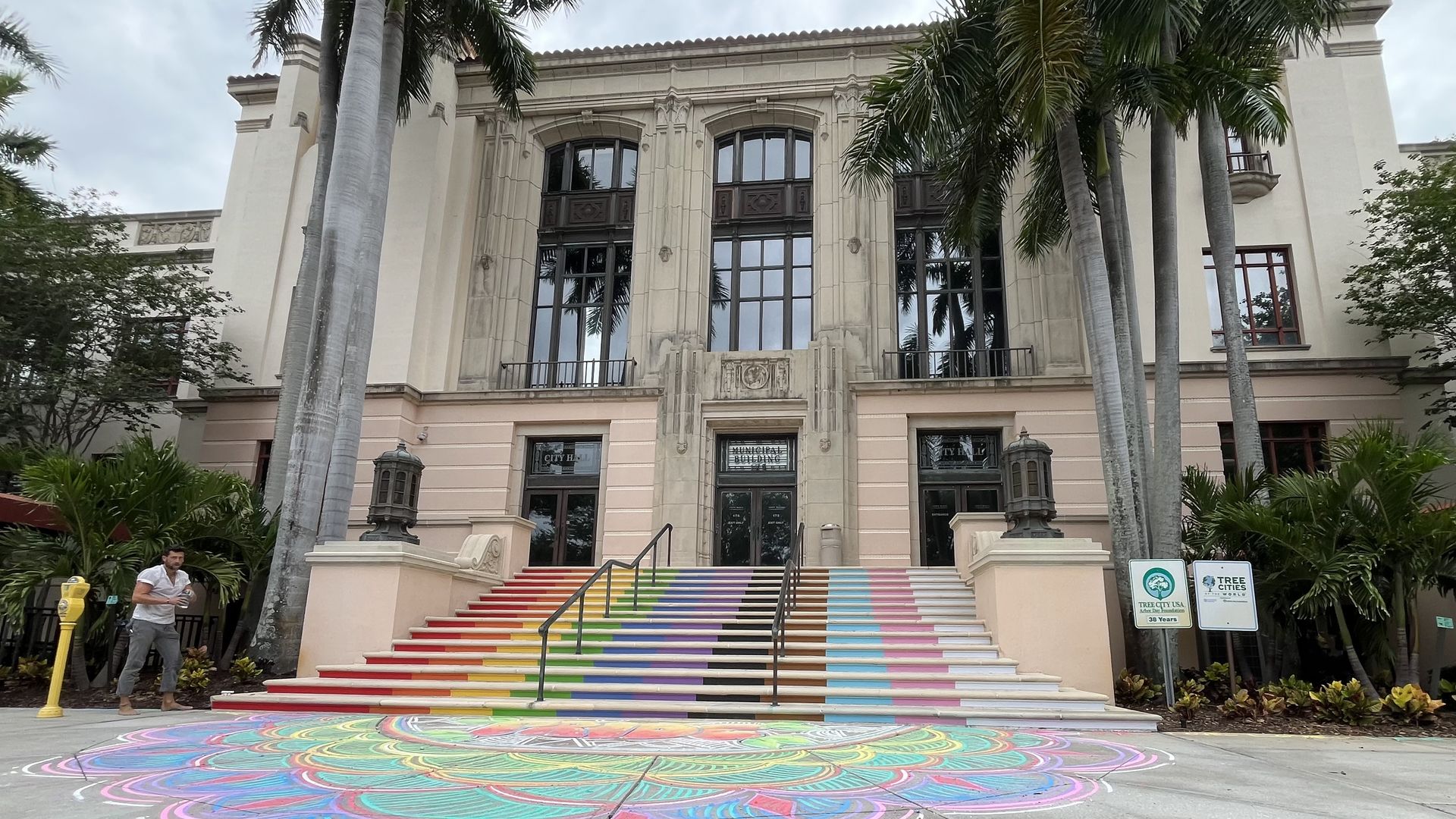 Front view of a beige neoclassical building framed by tall palm trees. Rainbow-striped stairs lead to glass doors; a large colorful chalk circle covers the pavement.