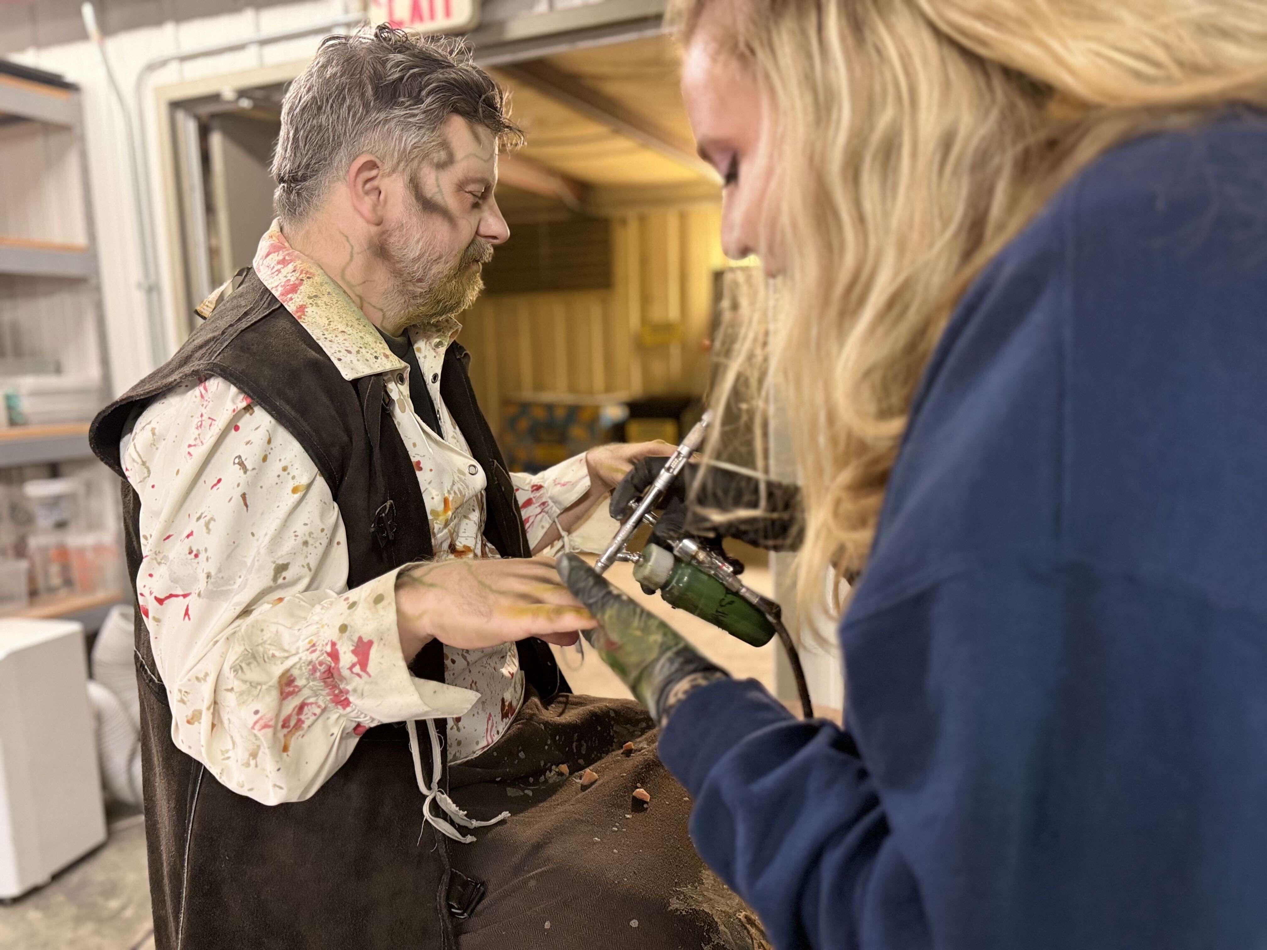 Bearded man in a paint-splattered shirt and vest in a workshop receives a tool from a blonde person in a blue-sleeved top; shelves and equipment visible in the background.