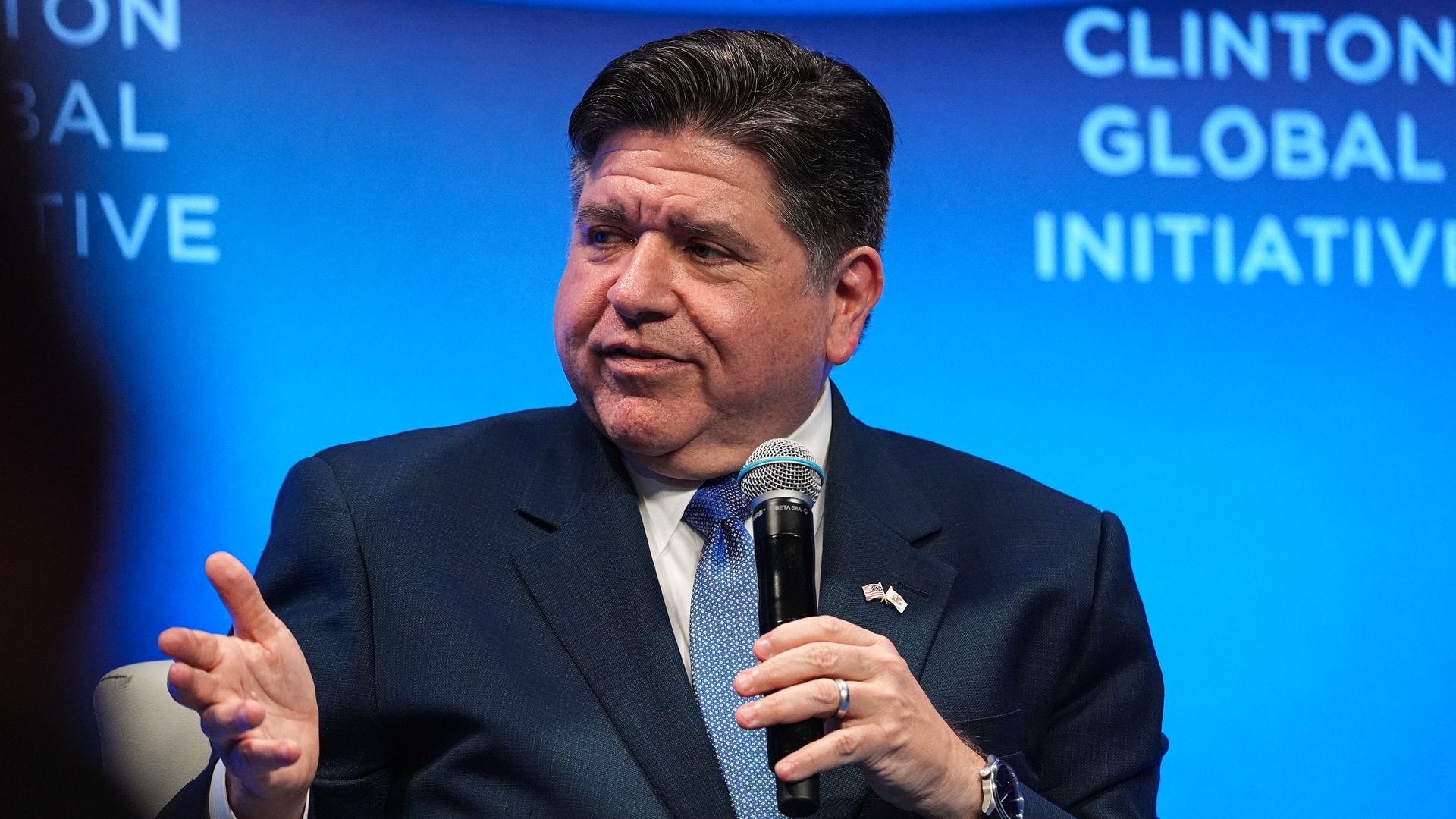 Man in a dark suit and blue tie speaks into a handheld microphone on a blue stage with the words "Clinton Global Initiative" visible in the background.