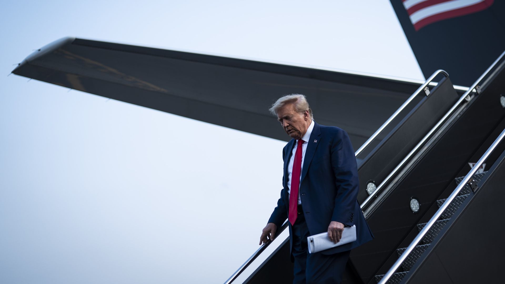 Former President Trump disembarking his airplane, wearing a dark blue suit, white shirt and red tie and holding papers.