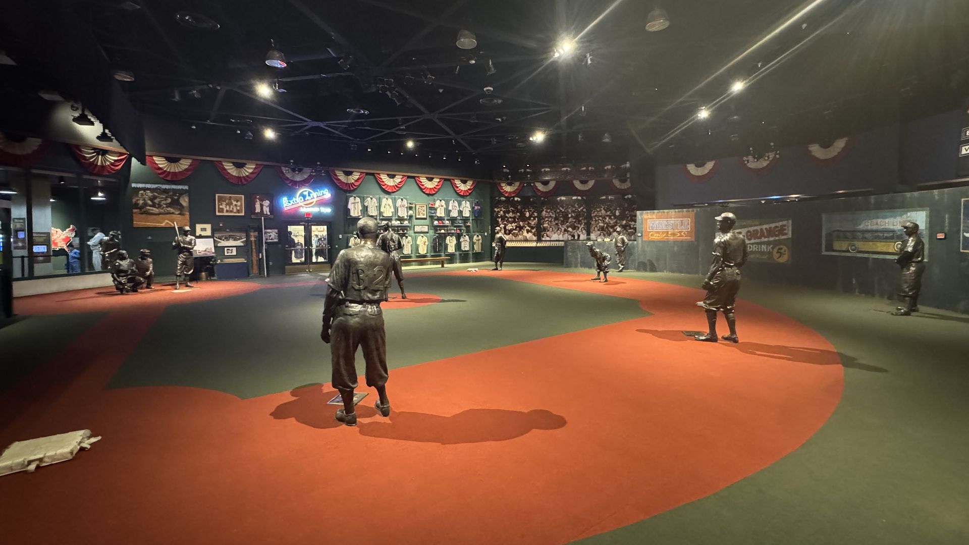 Indoor baseball museum scene with life-size metallic statues of players on a red clay infield, dark walls lined with vintage posters, bunting, and bright overhead spotlights.
