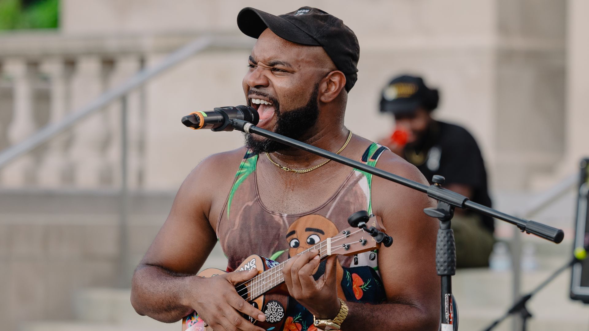 A man with a black cap sings into a microphone while strumming a small ukulele. He wears a tropical-print tank top, a gold chain, and a watch; a blurred background shows another person.