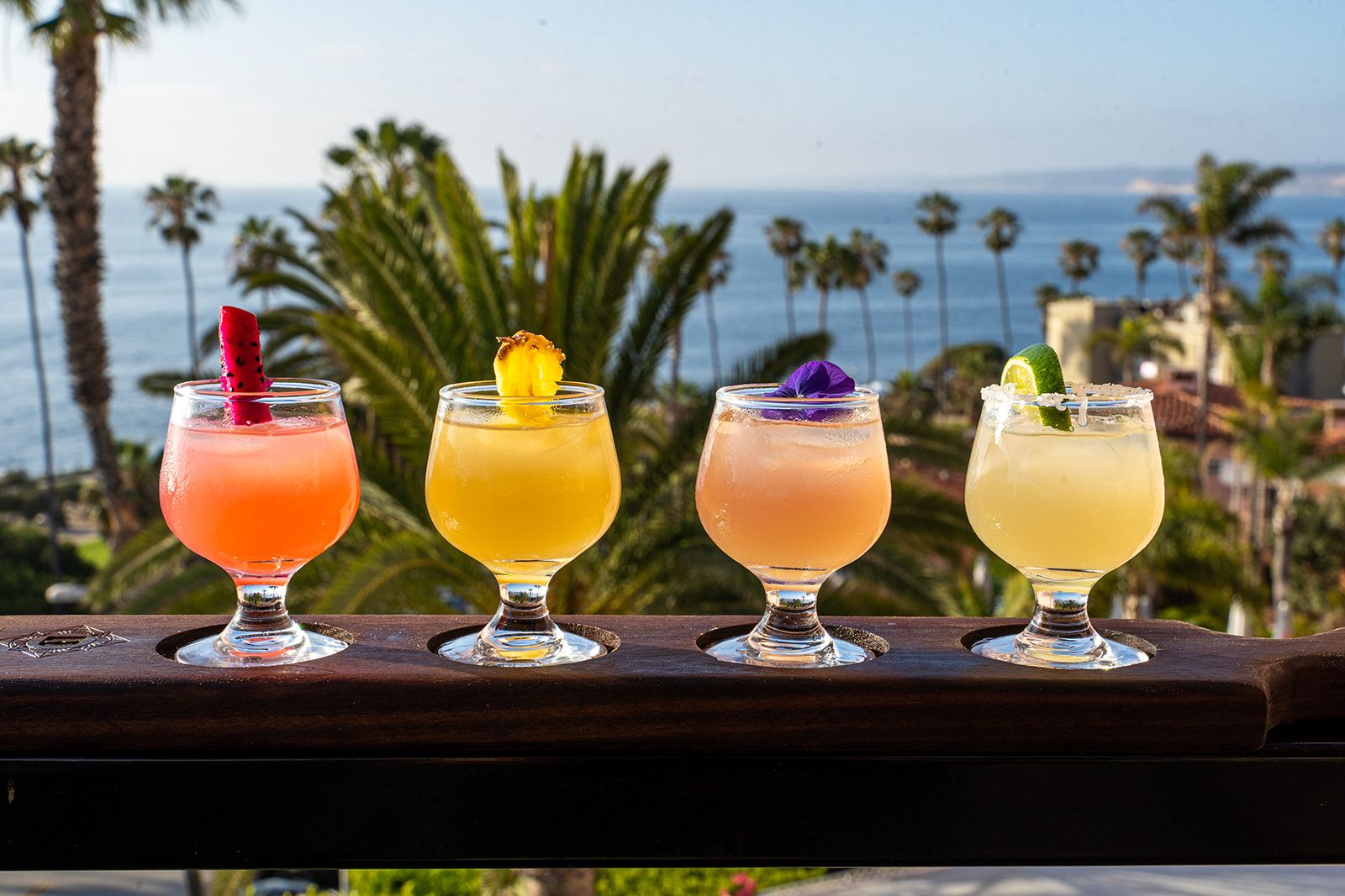 Four bright-colored cocktails with fruit garnishes sit on a wooden holder on a rooftop patio overlooking the ocean and palm trees.