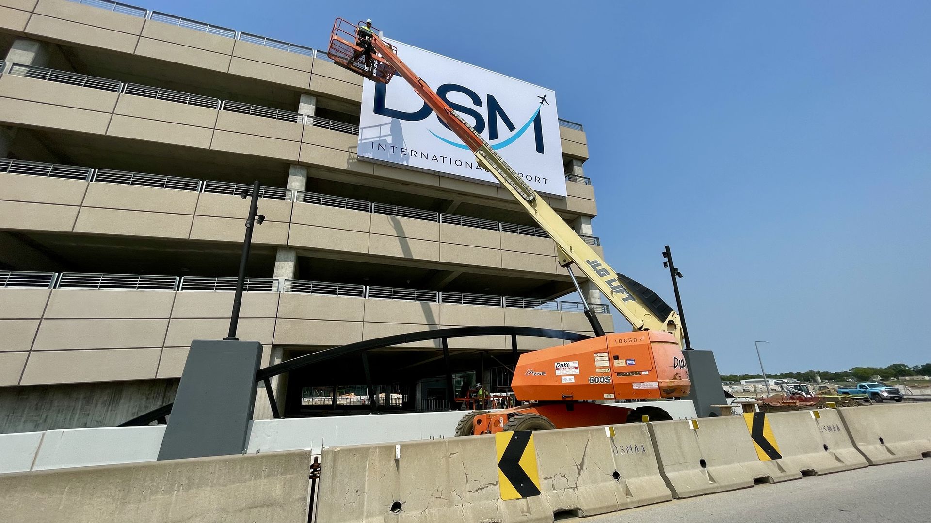 Orange and yellow boom lift with worker installing or maintaining a large DSM International Airport sign on a beige concrete parking garage under clear blue sky.