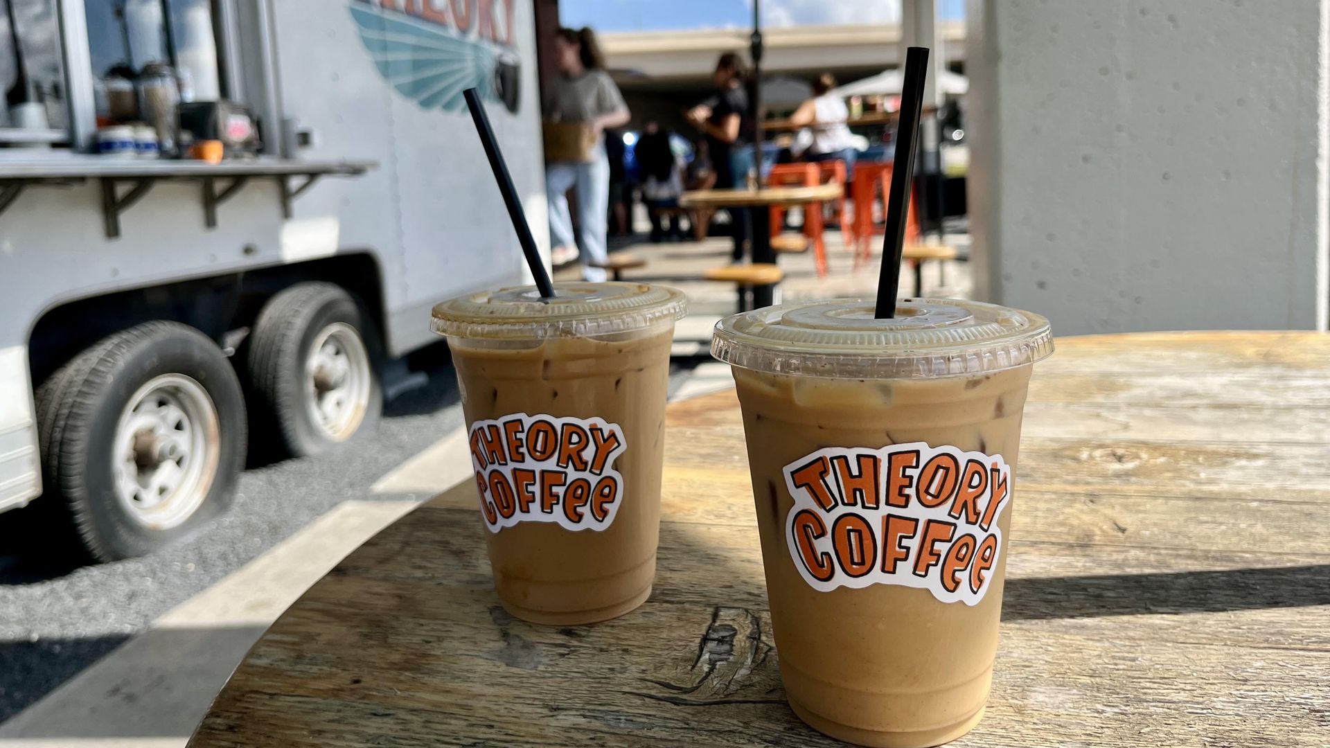 Two iced coffee drinks with black straws on a wooden table; blurred food truck and turquoise umbrella in the background, with people sitting at outdoor tables under a blue sky.